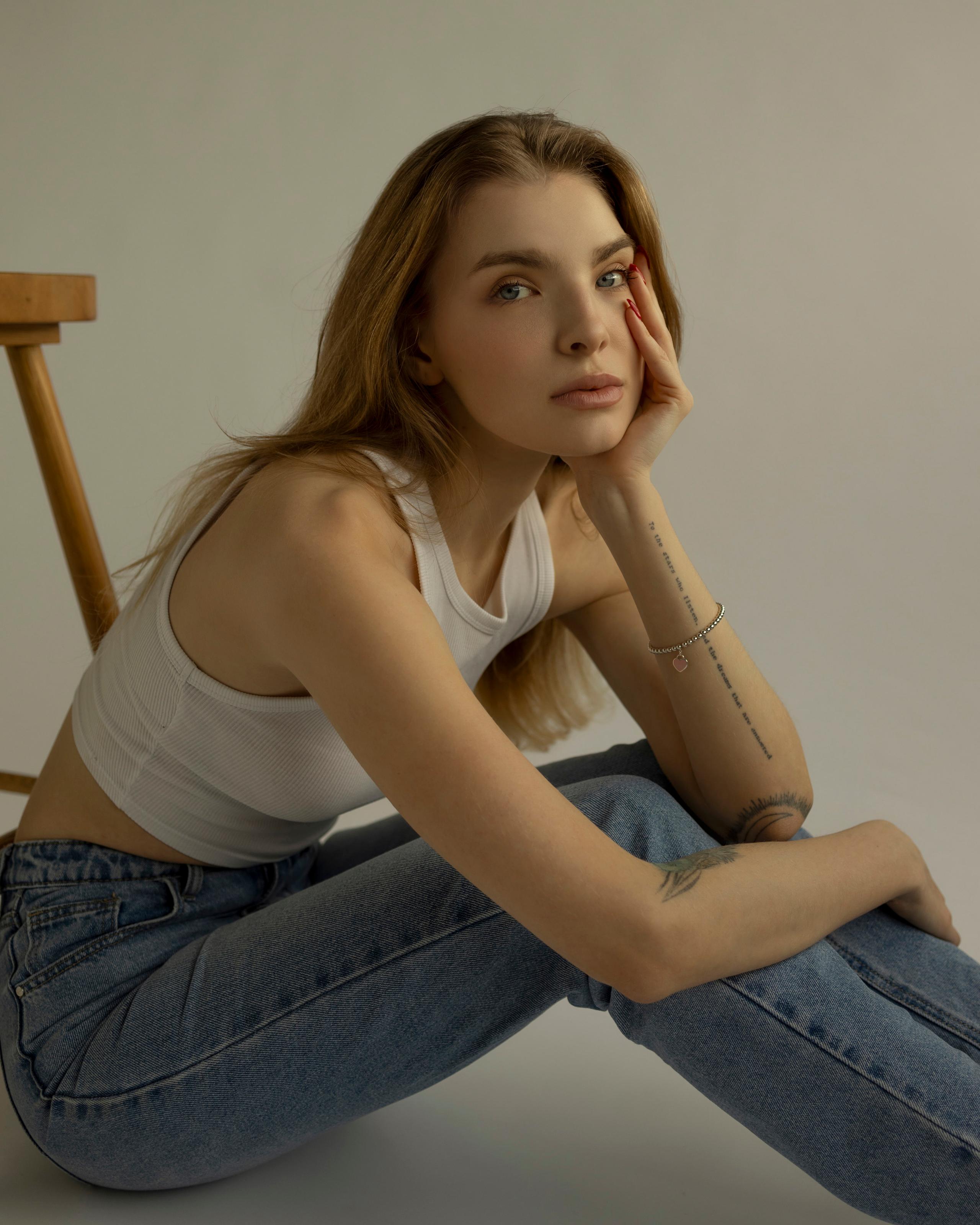 Auburn-haired woman in white tank and jeans, hand to face, seated by wooden chair, grey studio.