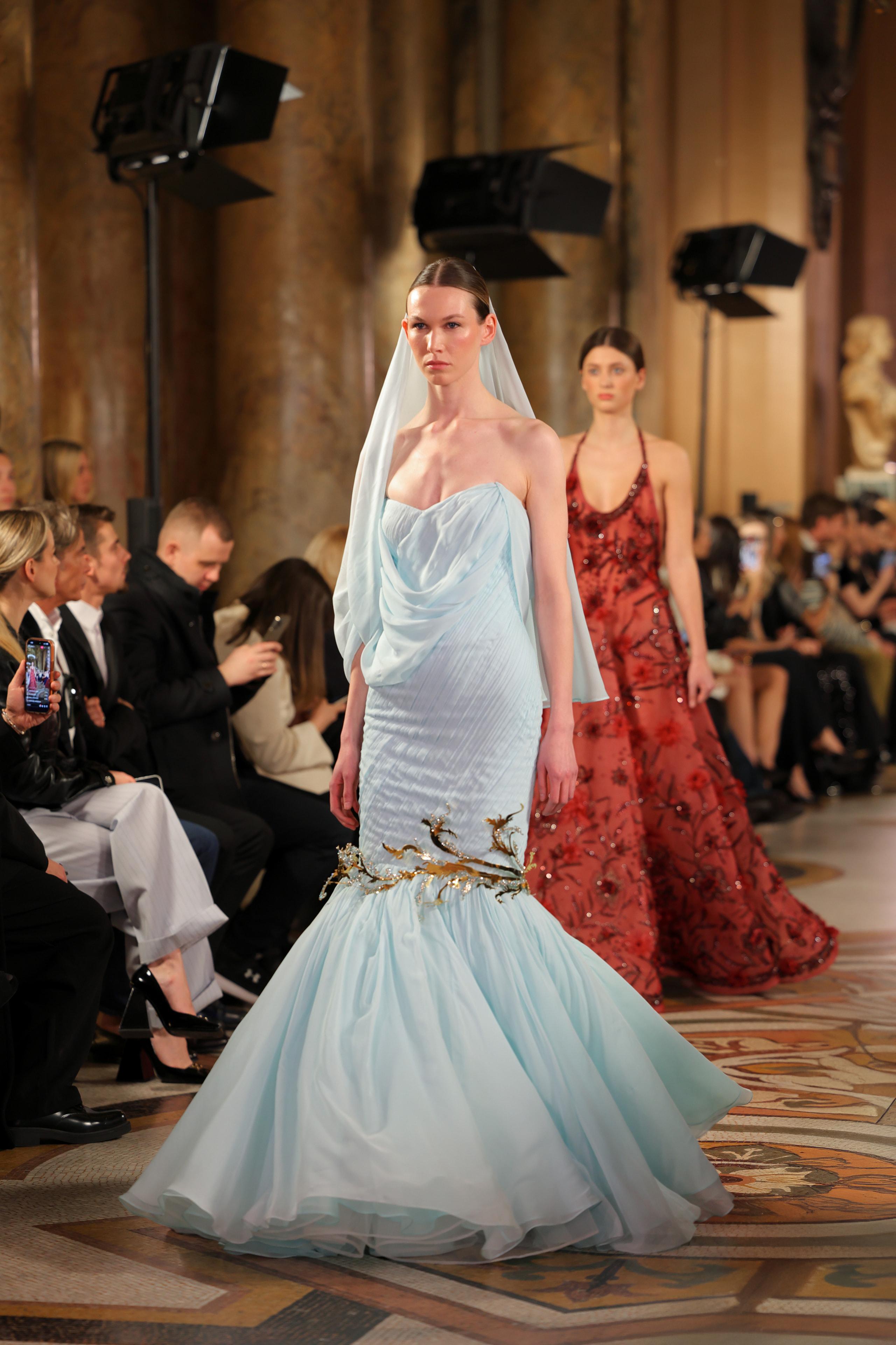 Model in pale blue strapless mermaid gown and veil leading second model on runway