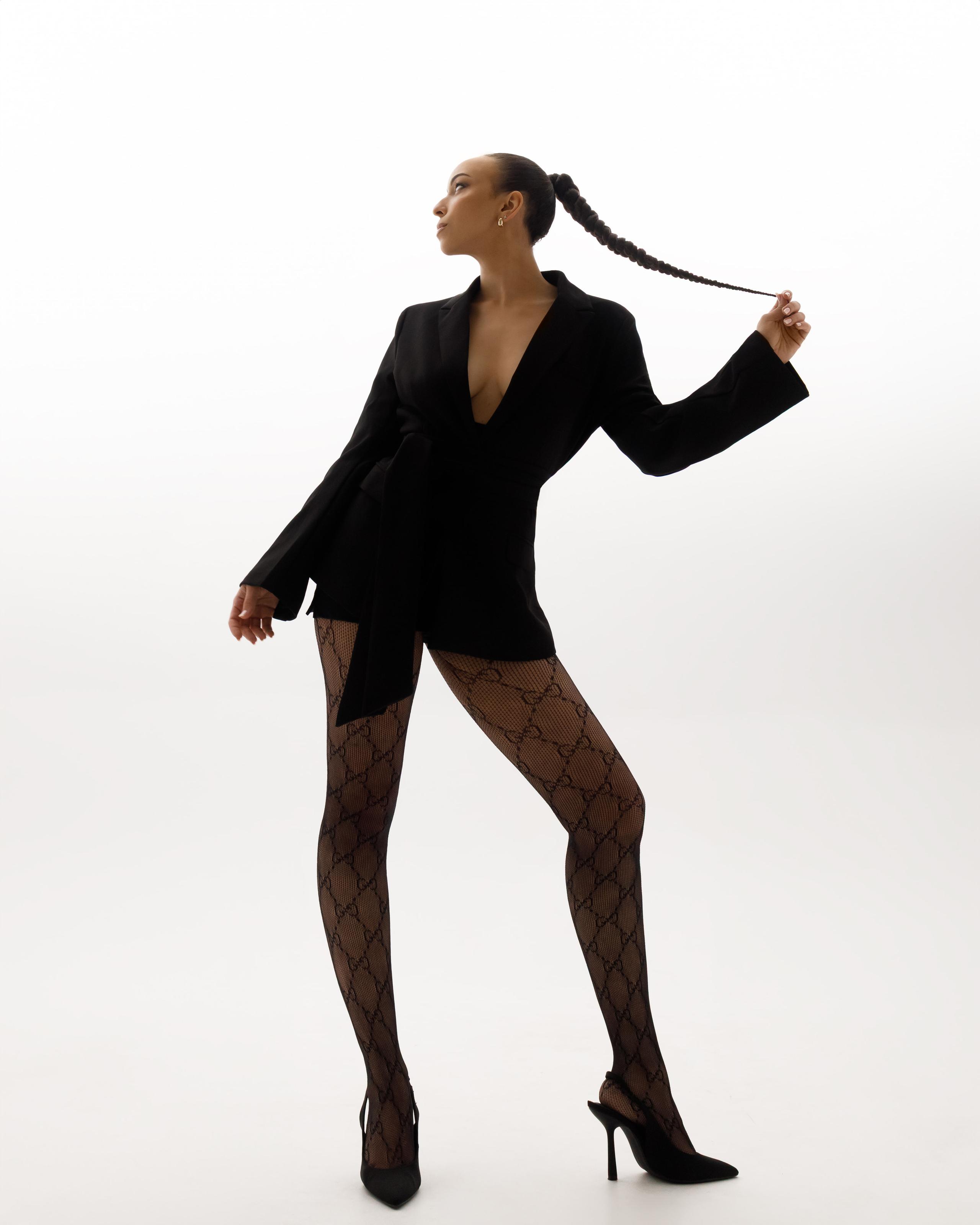 Full-length model in black blazer and monogram tights, pulling long braid, on white backdrop.