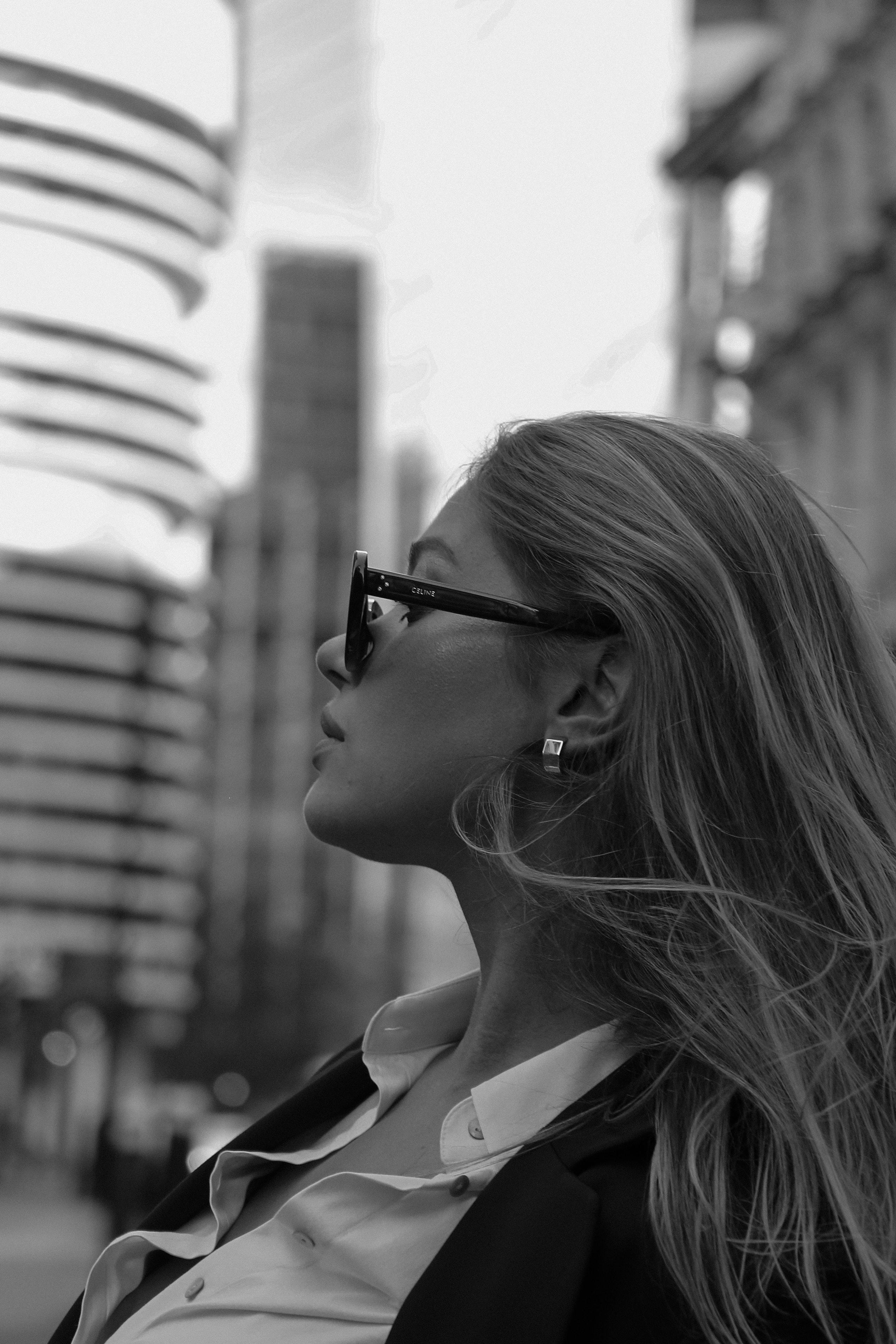 Black-and-white profile of woman in Celine sunglasses gazing upward against blurred London skyscrapers