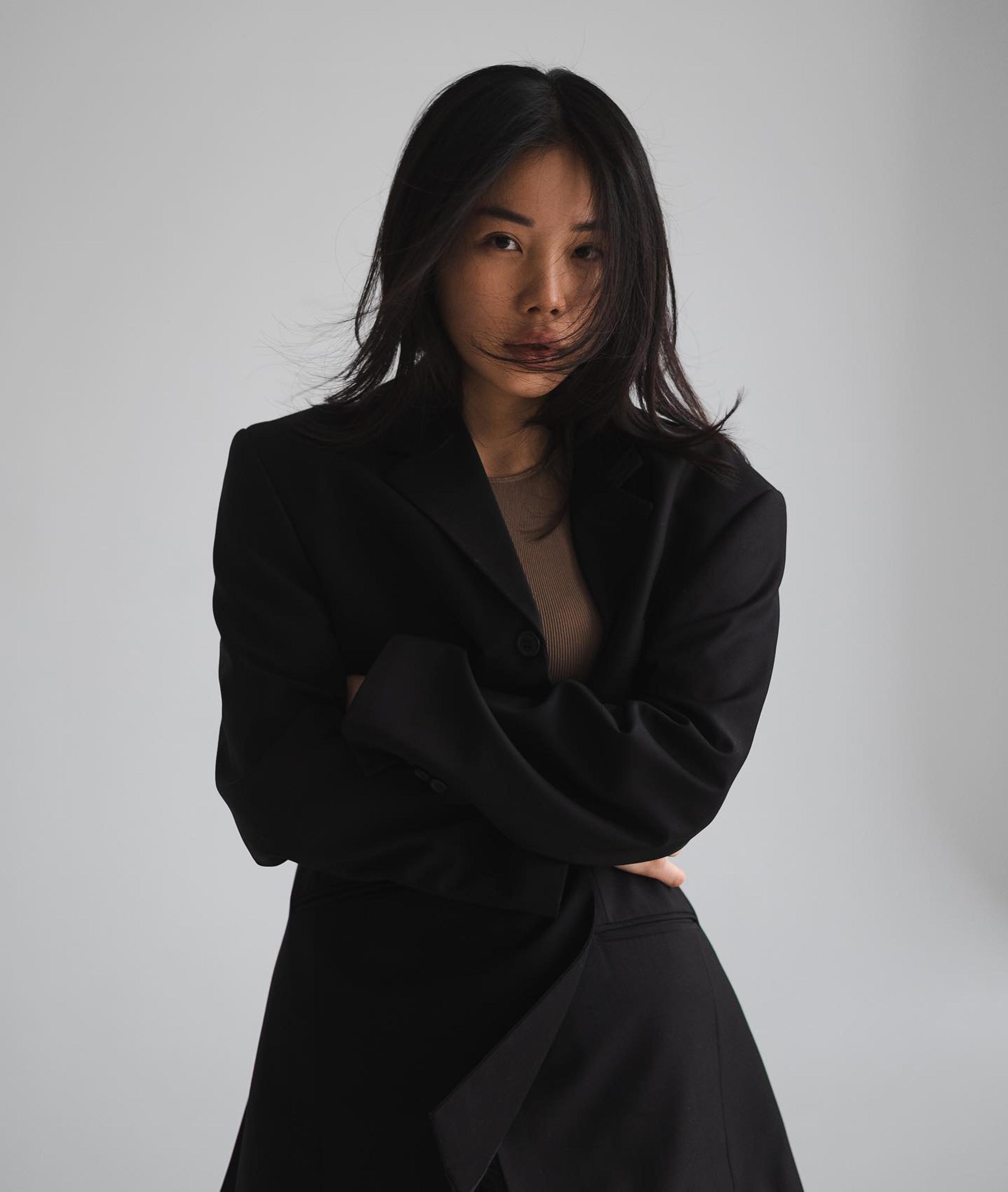 Dark-haired woman in black blazer over sheer brown top, arms crossed, windblown hair, grey backdrop