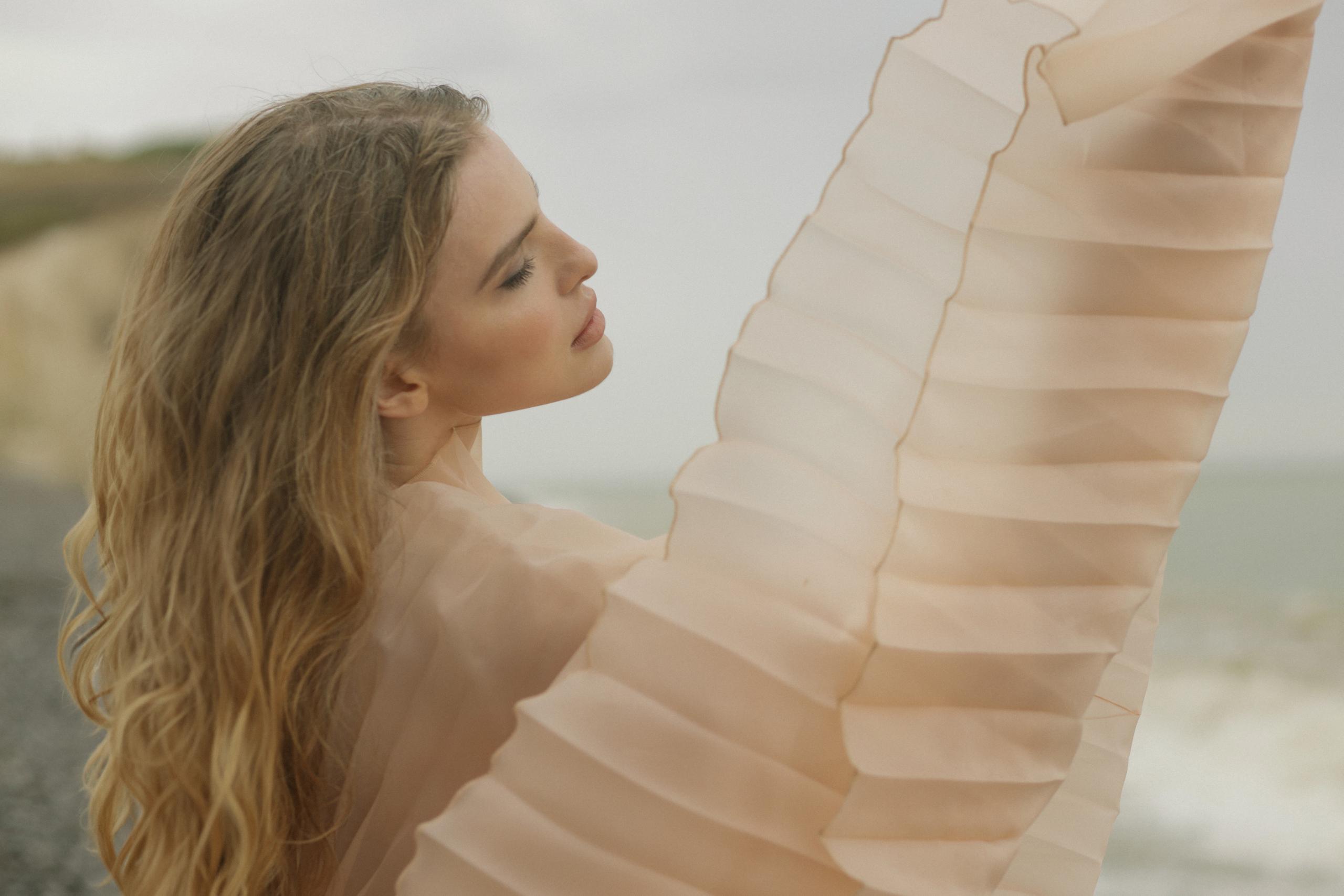 Profile of woman with eyes closed, sheer peach pleated sleeve lifted dramatically against pale coastal sky