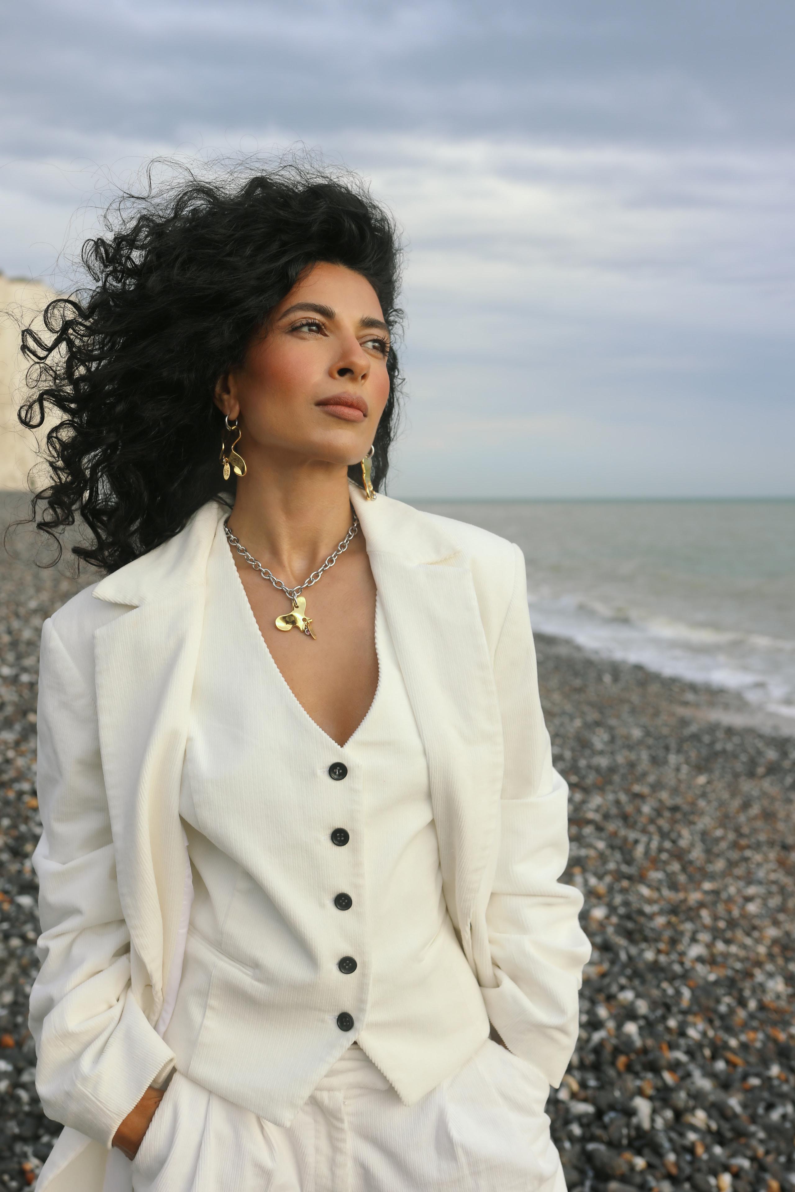 Woman with voluminous dark curls in cream suit and gold jewellery gazing off across grey sea