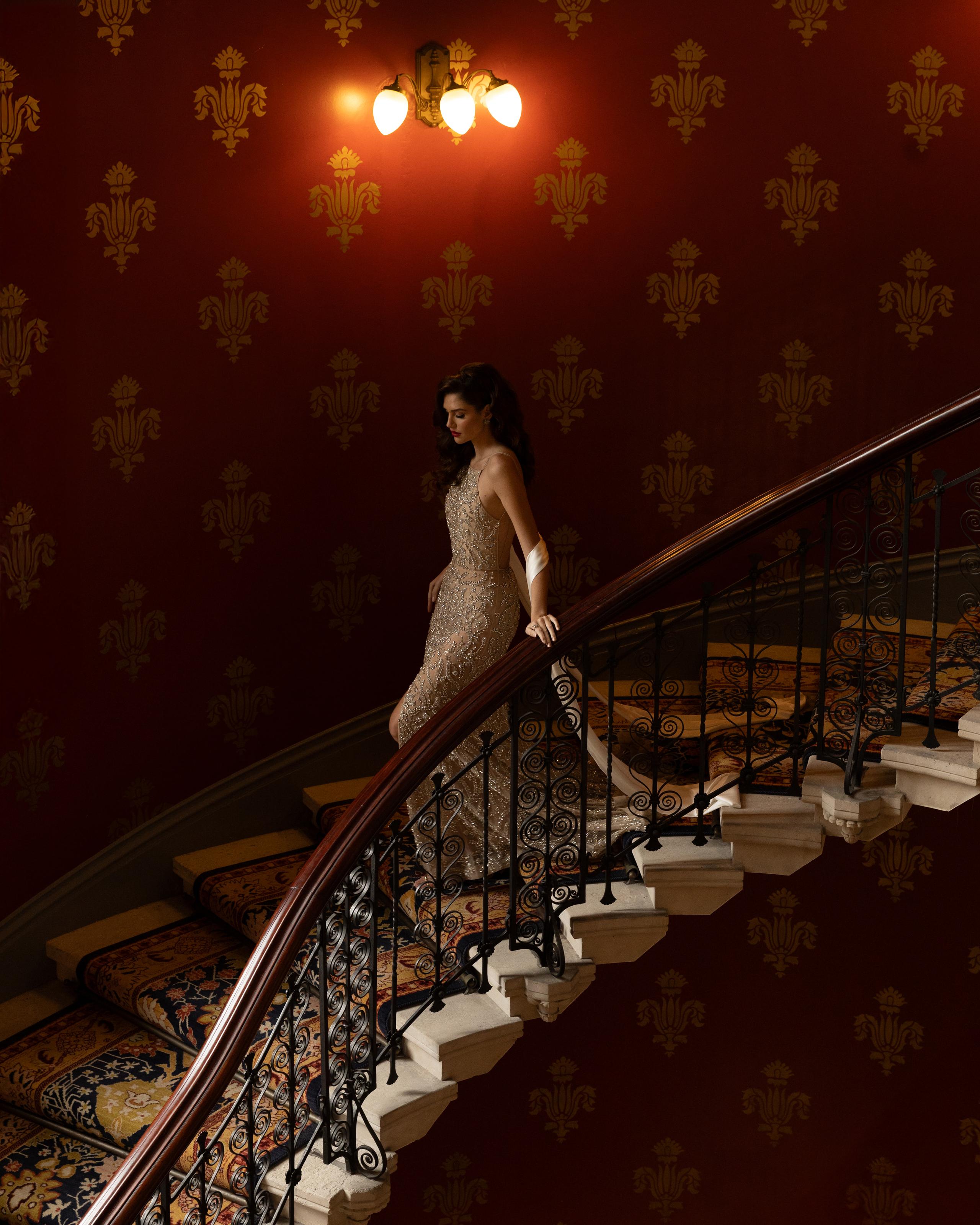 Model in beaded gown descending sweeping staircase against red damask wall and ornate carpet