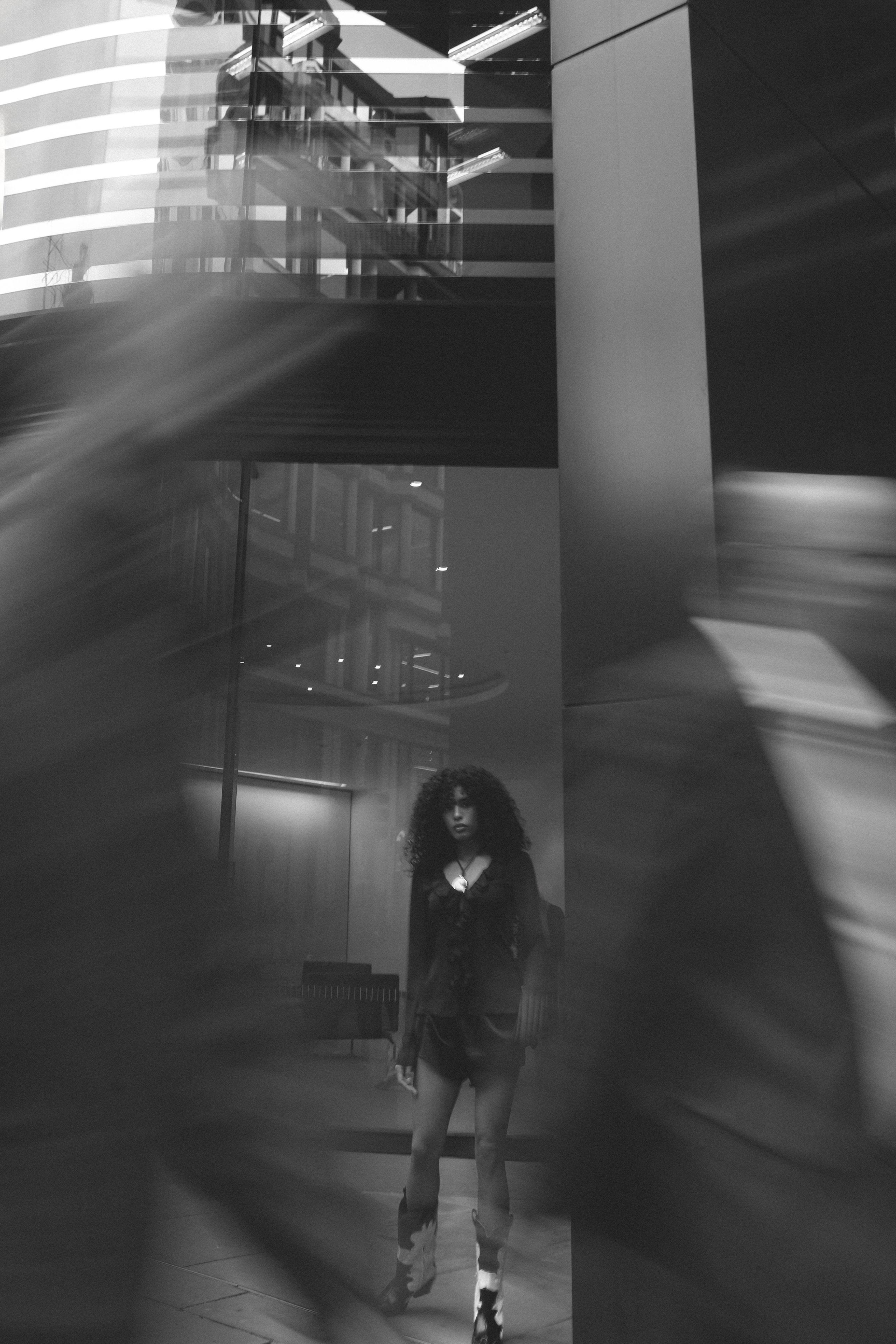 Model walks through a glass office atrium amid layered reflections in moody black and white