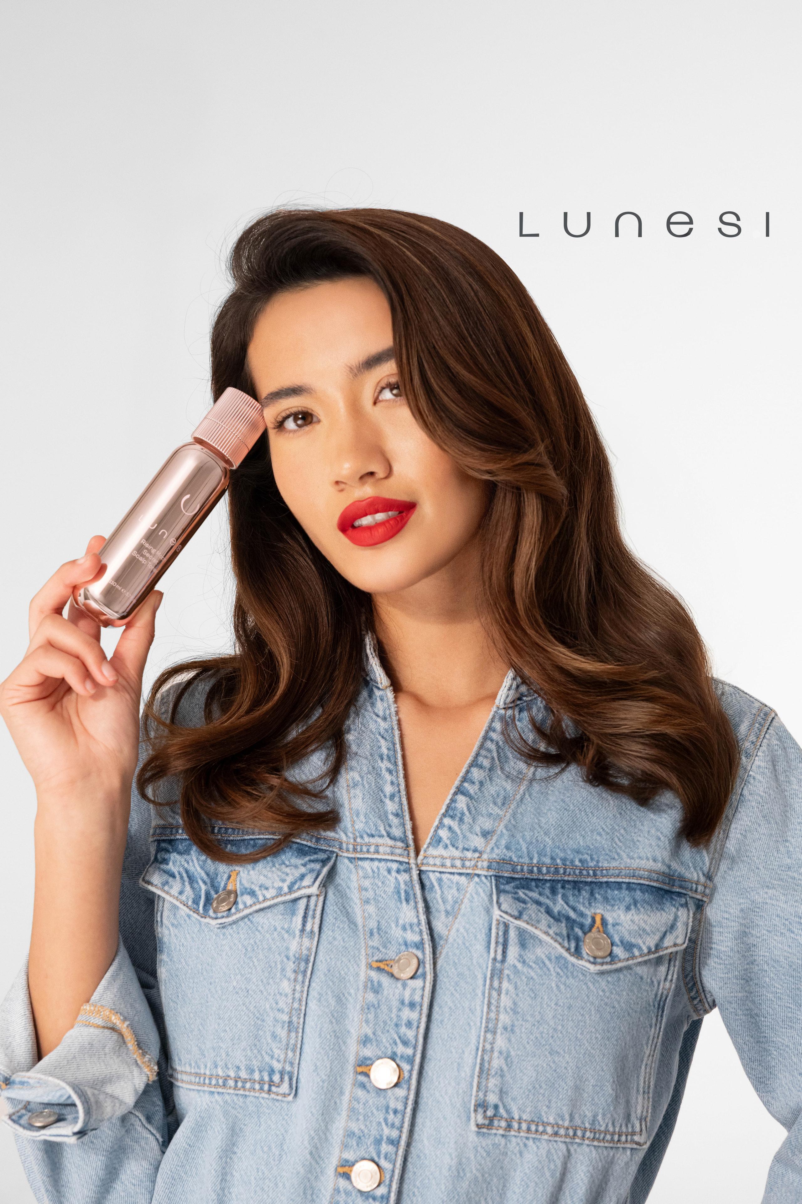 Brunette model in a denim jacket holds a rose-gold Lunesi serum bottle near her face, red lips