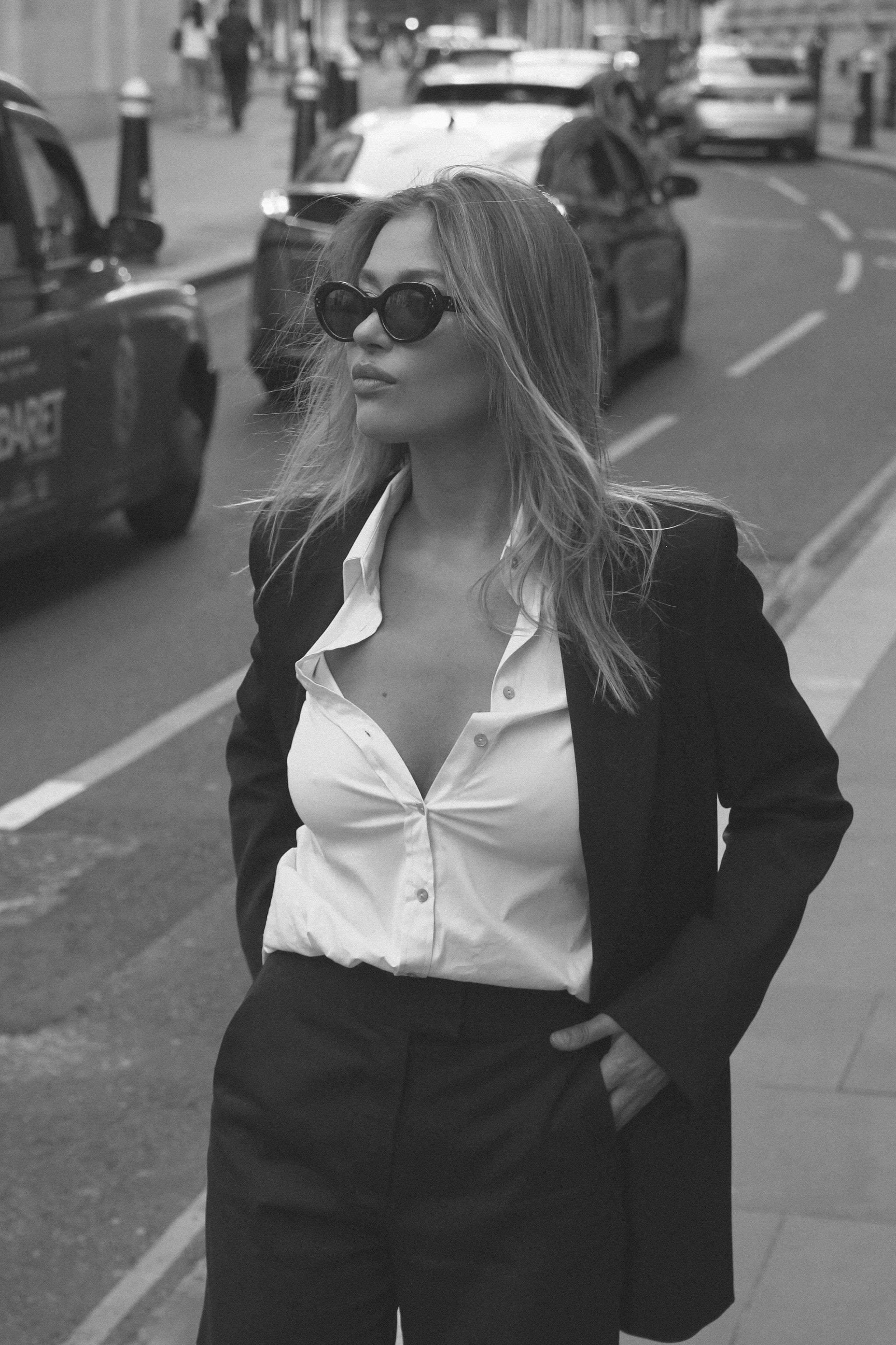 Black-and-white street portrait of woman in tailored blazer and white shirt, hand on hip, London