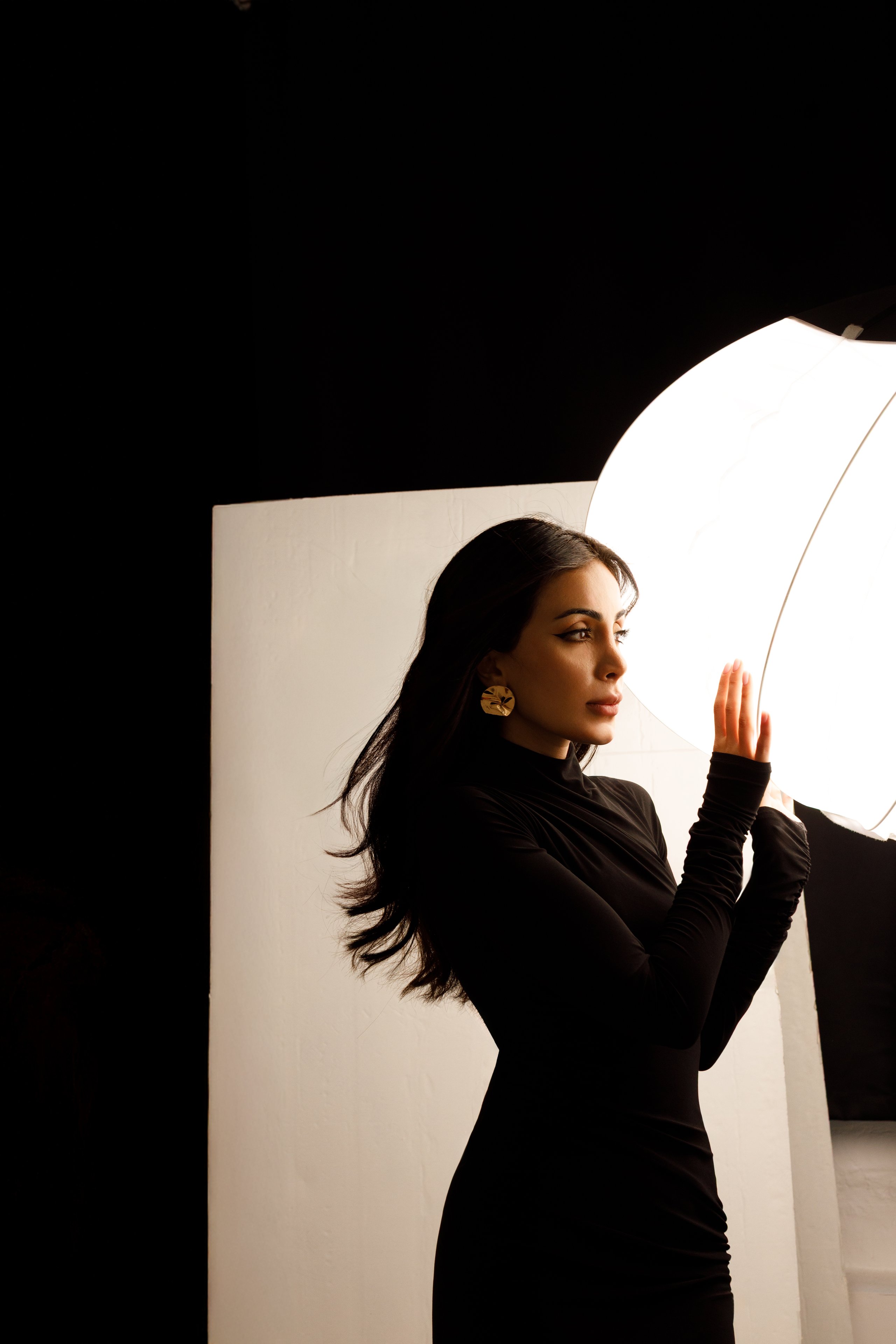 Model in a black dress raising a hand toward a glowing octabox light between takes in a darkened studio