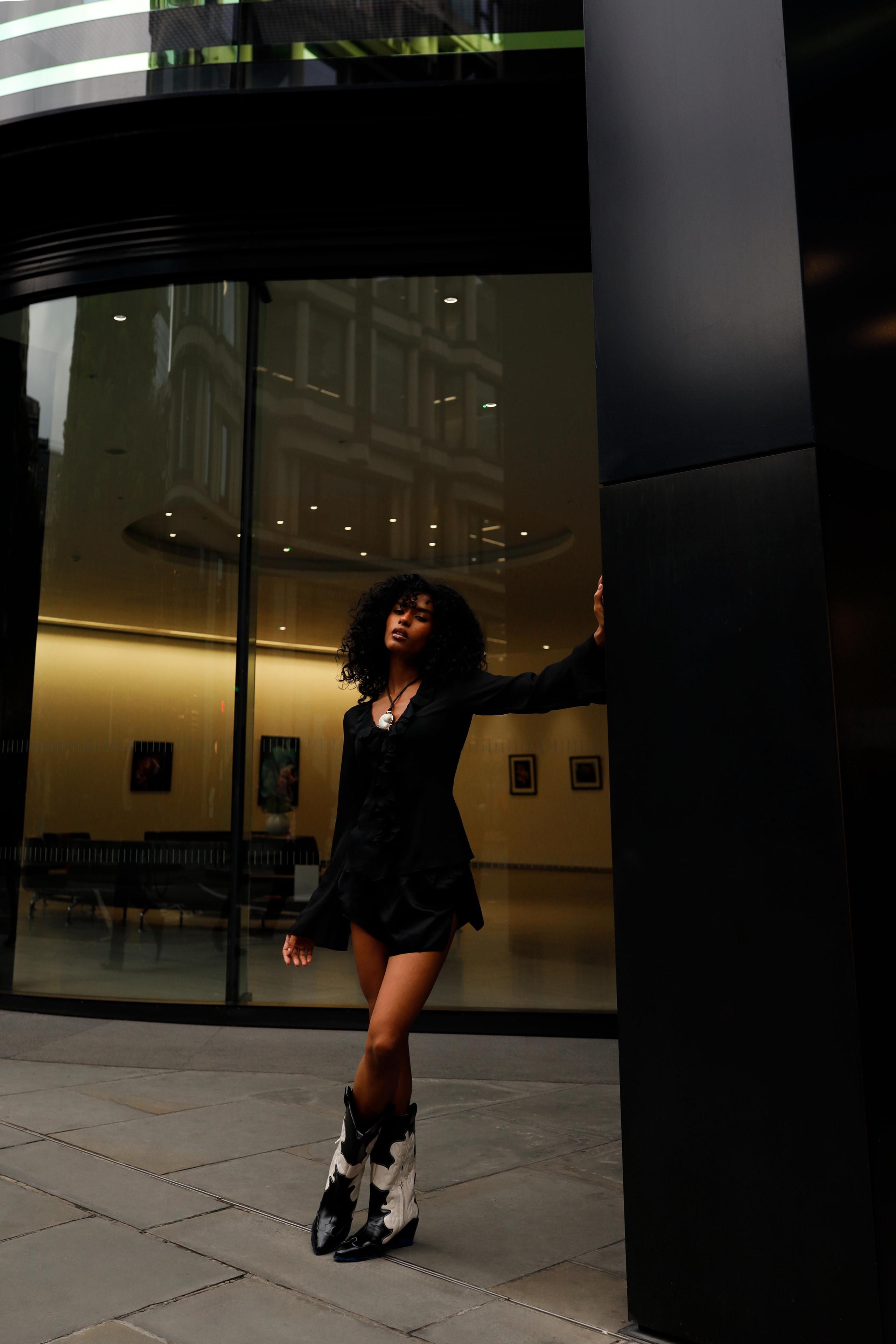 Model in black blazer dress leans against a column outside a warm-lit glass office lobby