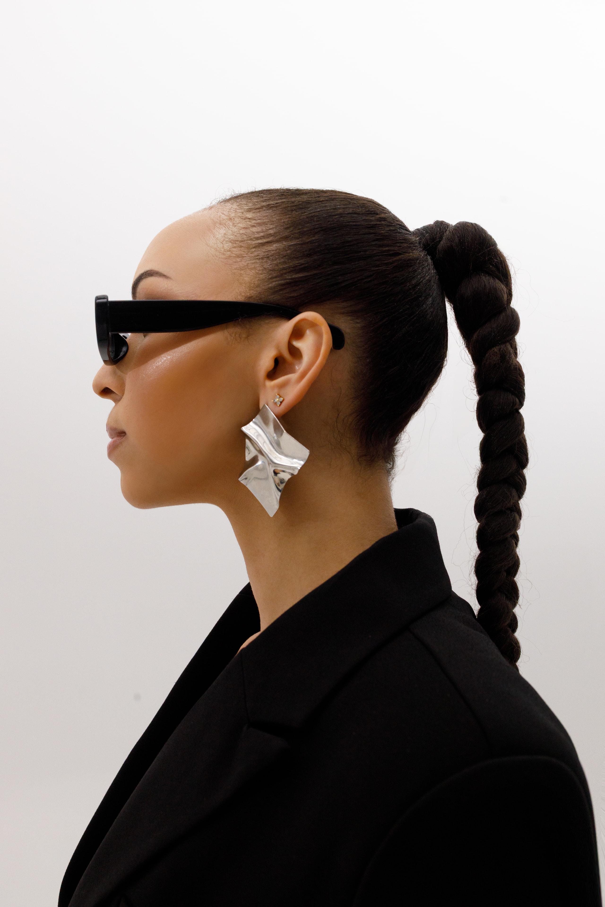 Profile of woman in black blazer and sunglasses with sculptural silver earring and braided ponytail