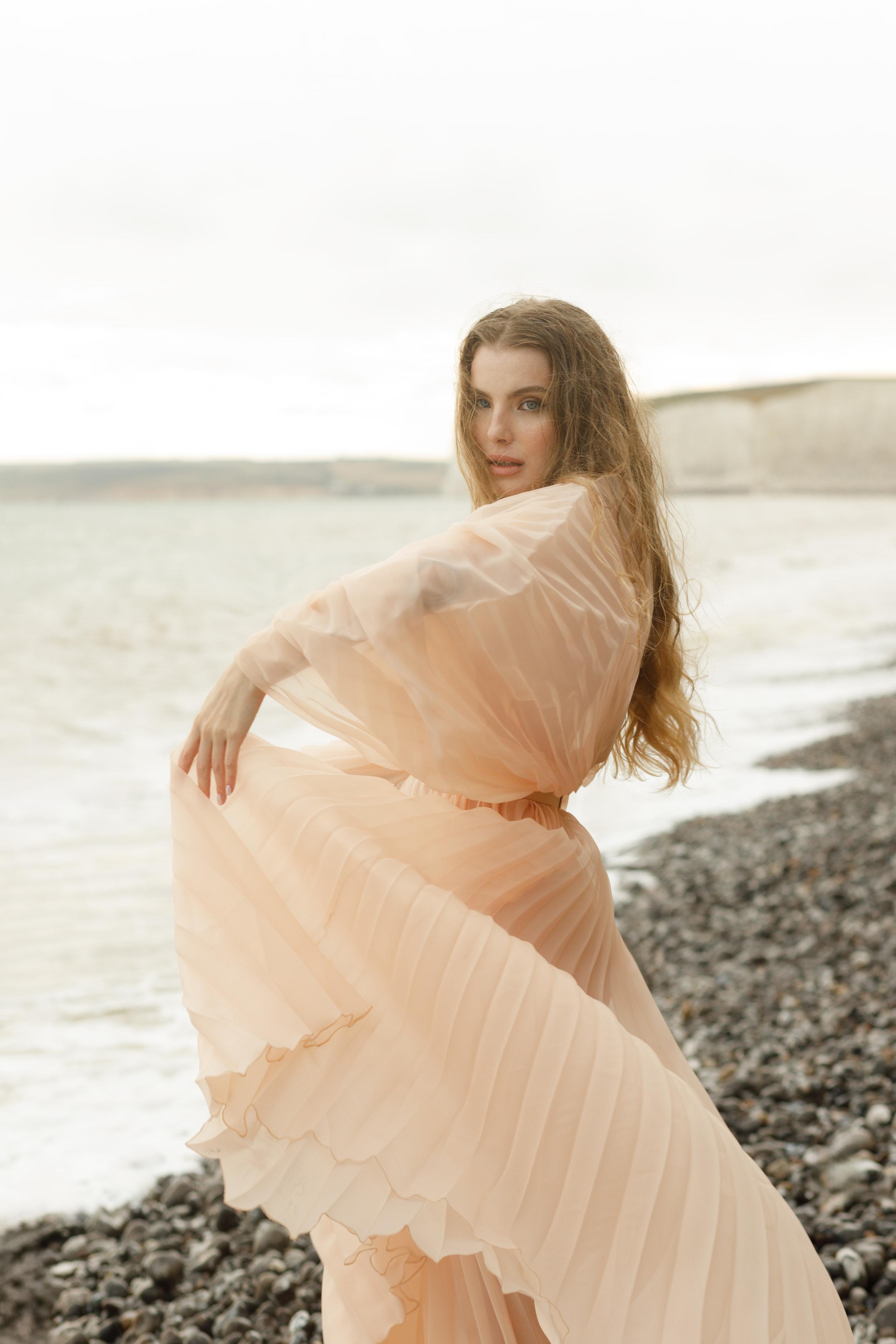 Woman turning over shoulder in flowing peach pleated gown on pebble beach, hair caught in wind