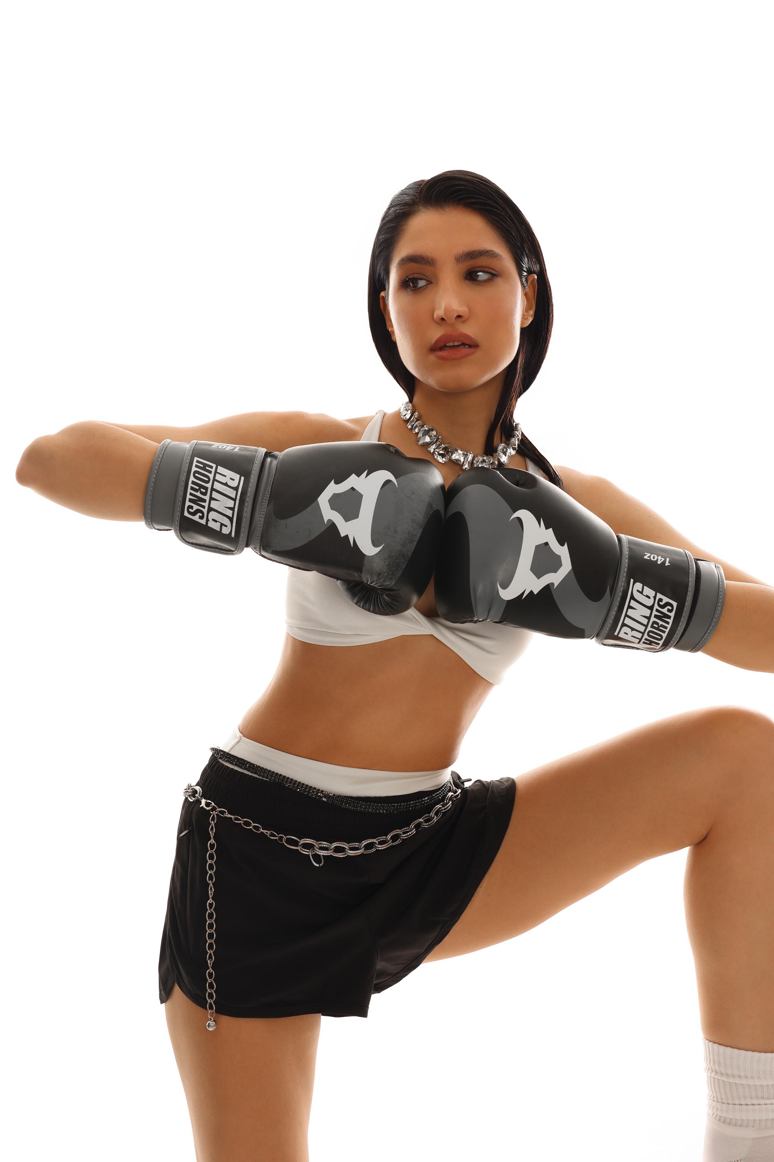 Woman in white crop top and chain-belt black skirt fists together in Ringhorns boxing gloves, white set