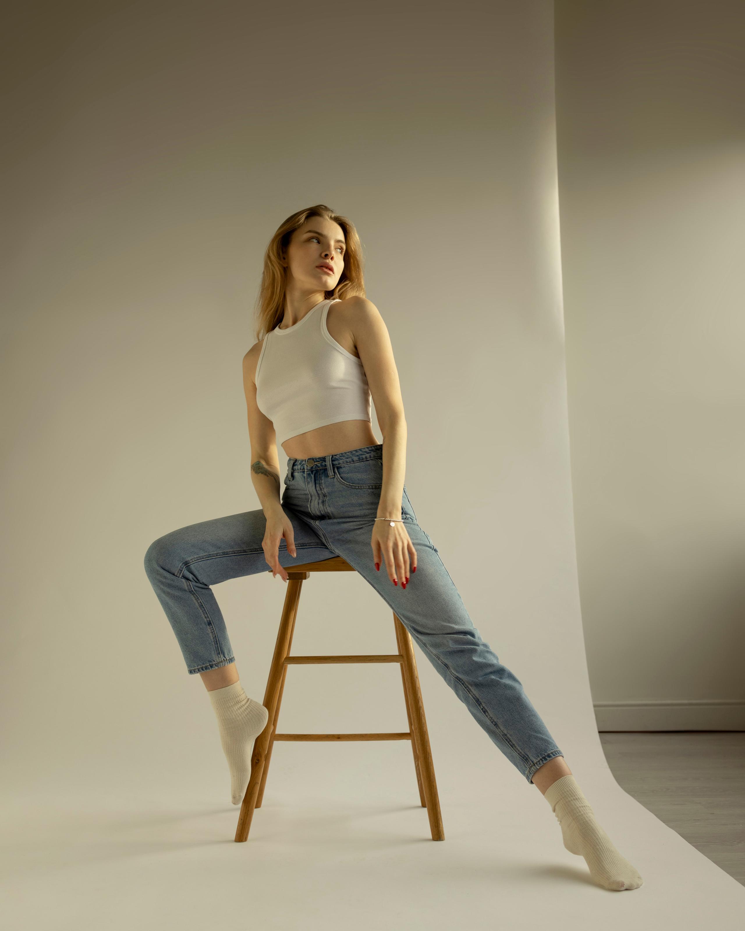 Woman in white tank and jeans perched on wooden stool, legs extended, soft beige studio sweep.