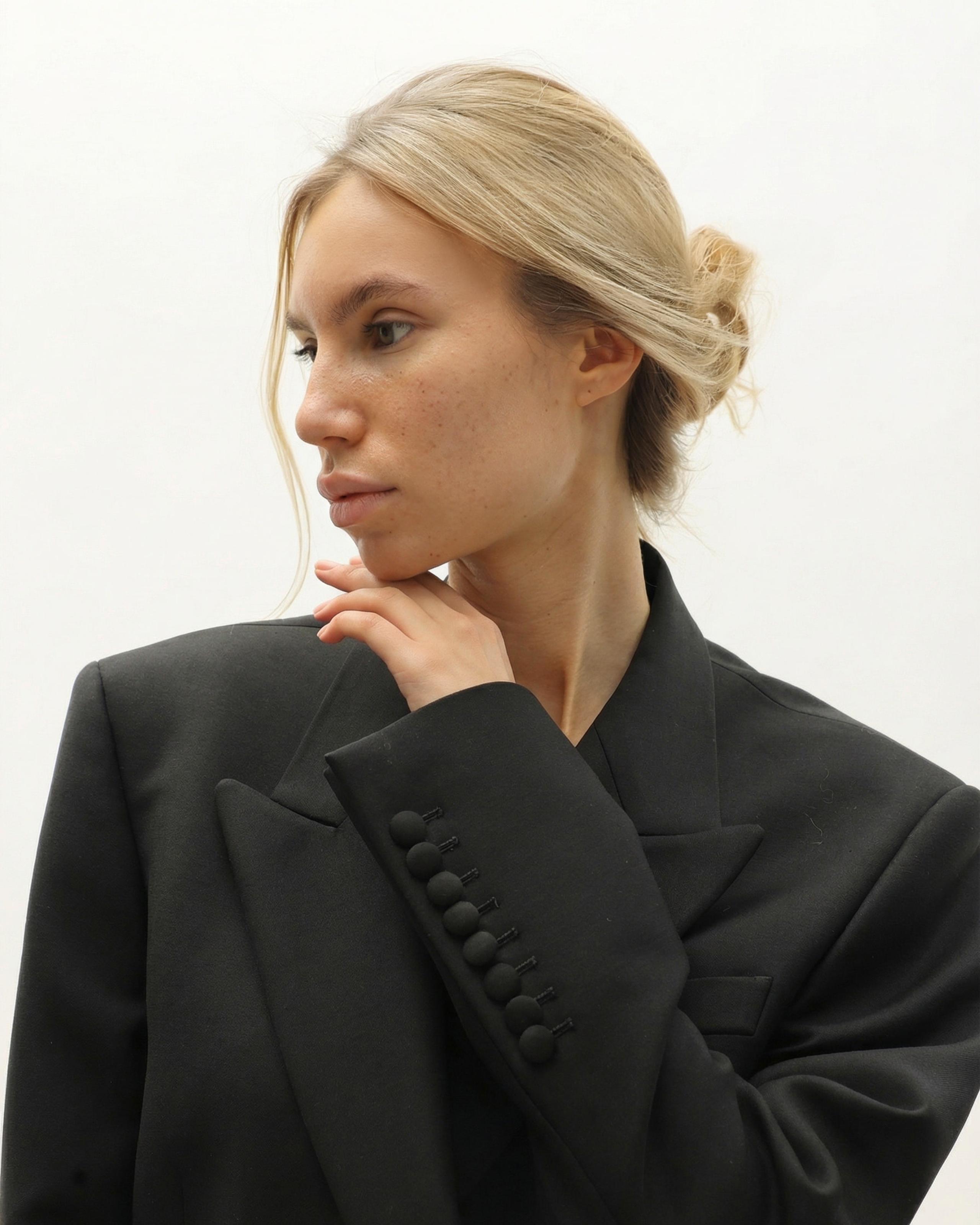 Blonde woman with freckles and a low bun in a black blazer, hand to chin, looking aside, white backdrop.