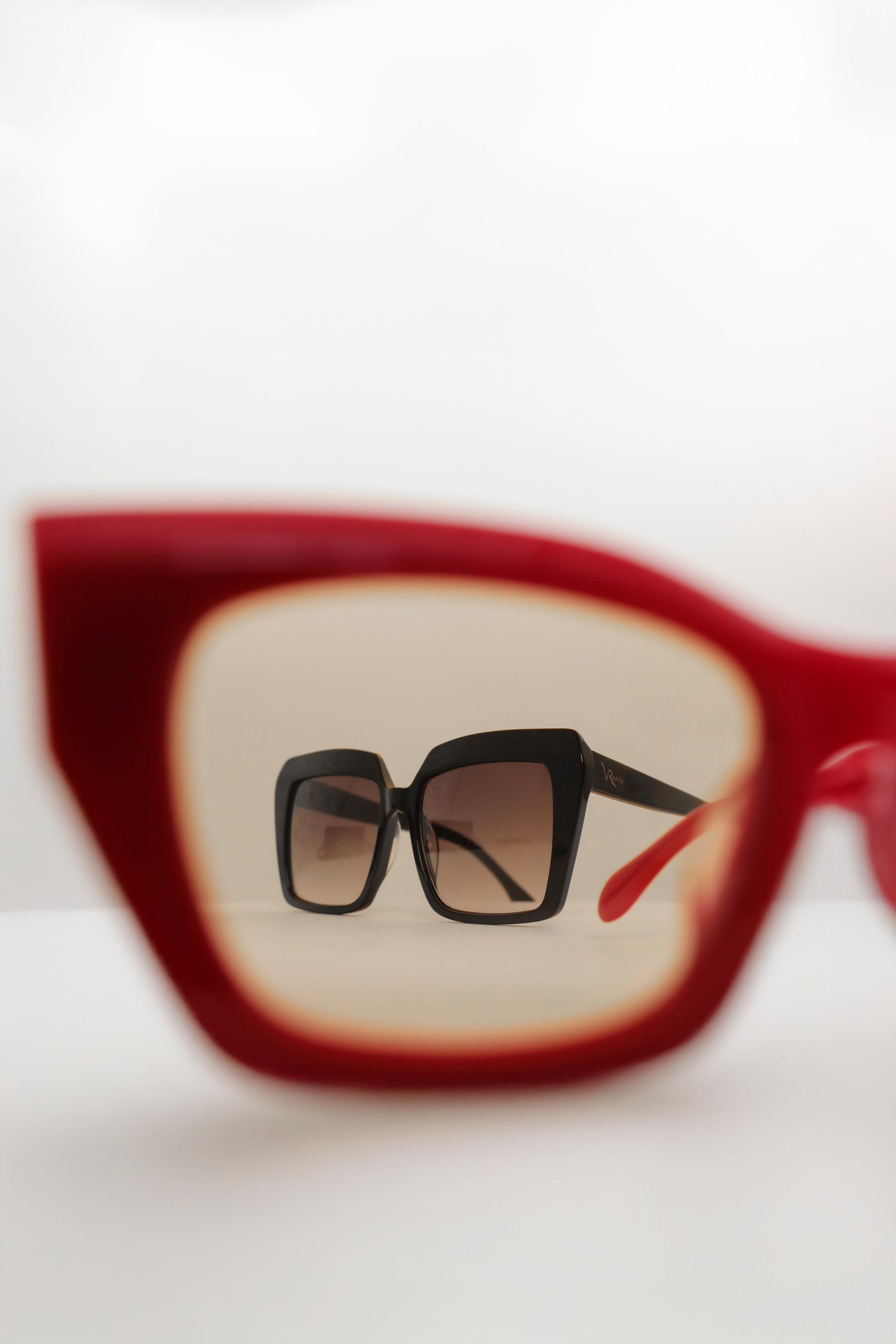 Black square sunglasses photographed through an out-of-focus red sunglasses frame on white surface