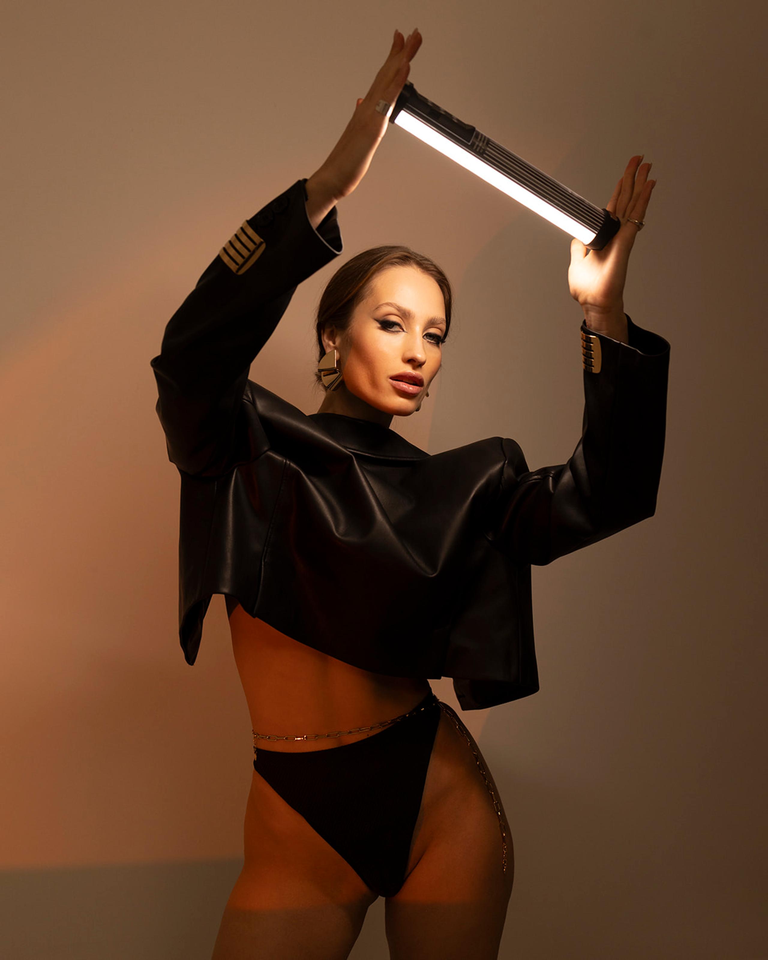 Model holding glowing LED tube overhead in leather jacket against warm taupe studio backdrop