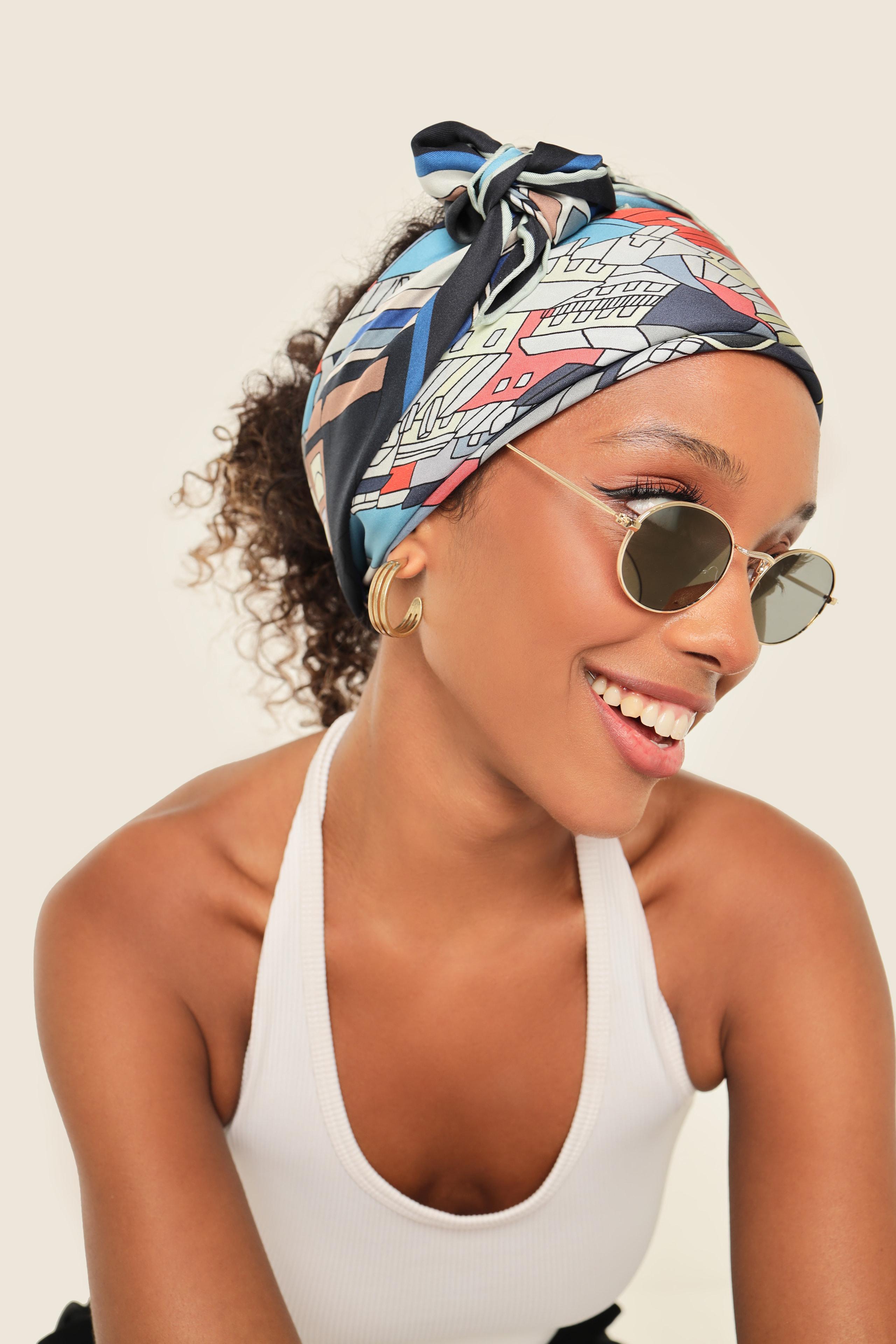 Smiling woman in a colorful printed headscarf, round sunglasses and white tank top.