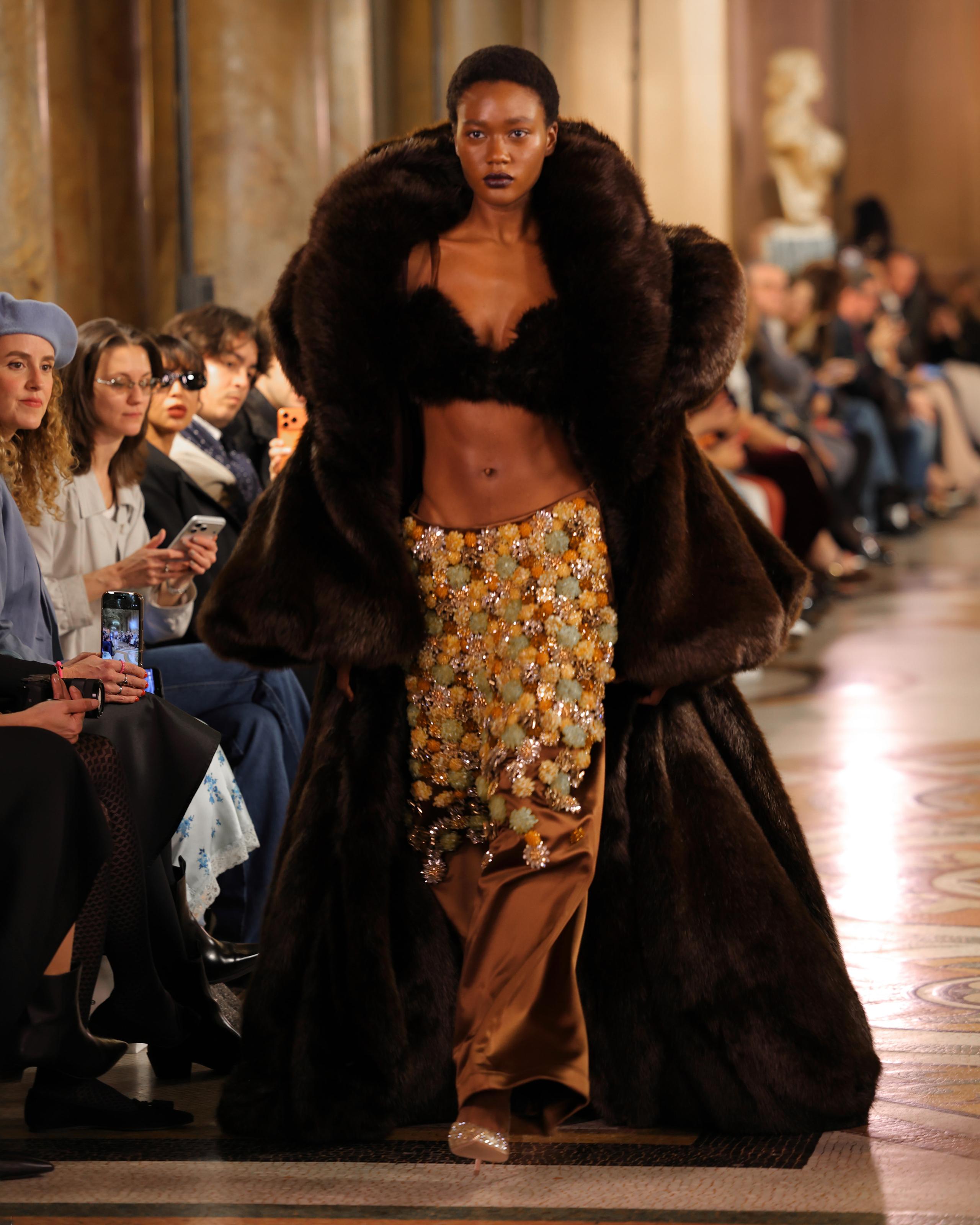 Model in oversized brown fur coat and beaded skirt on marble runway