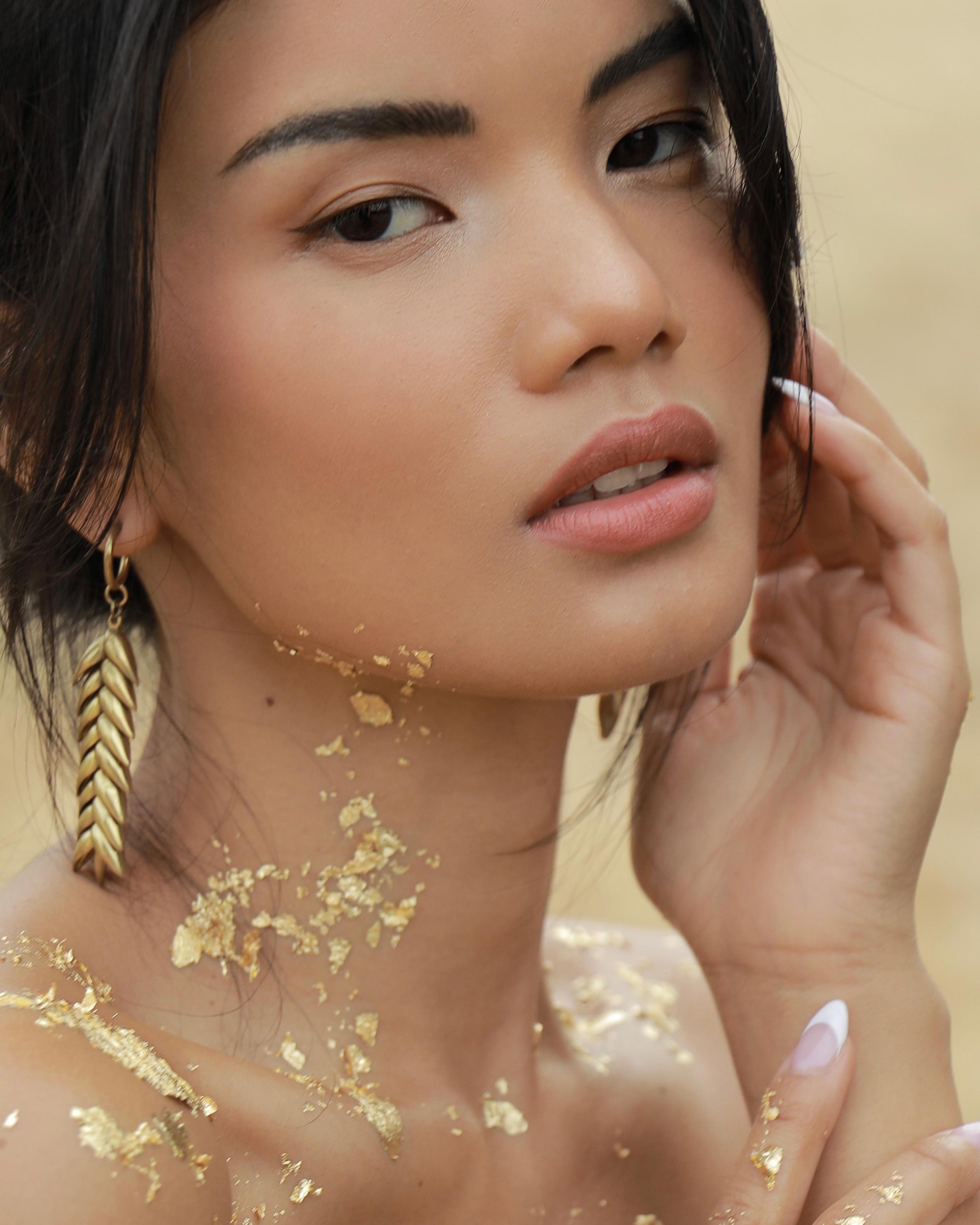 Close-up of woman with gold leaf flecks on skin, leaf earring, soft warm backdrop, bare shoulders