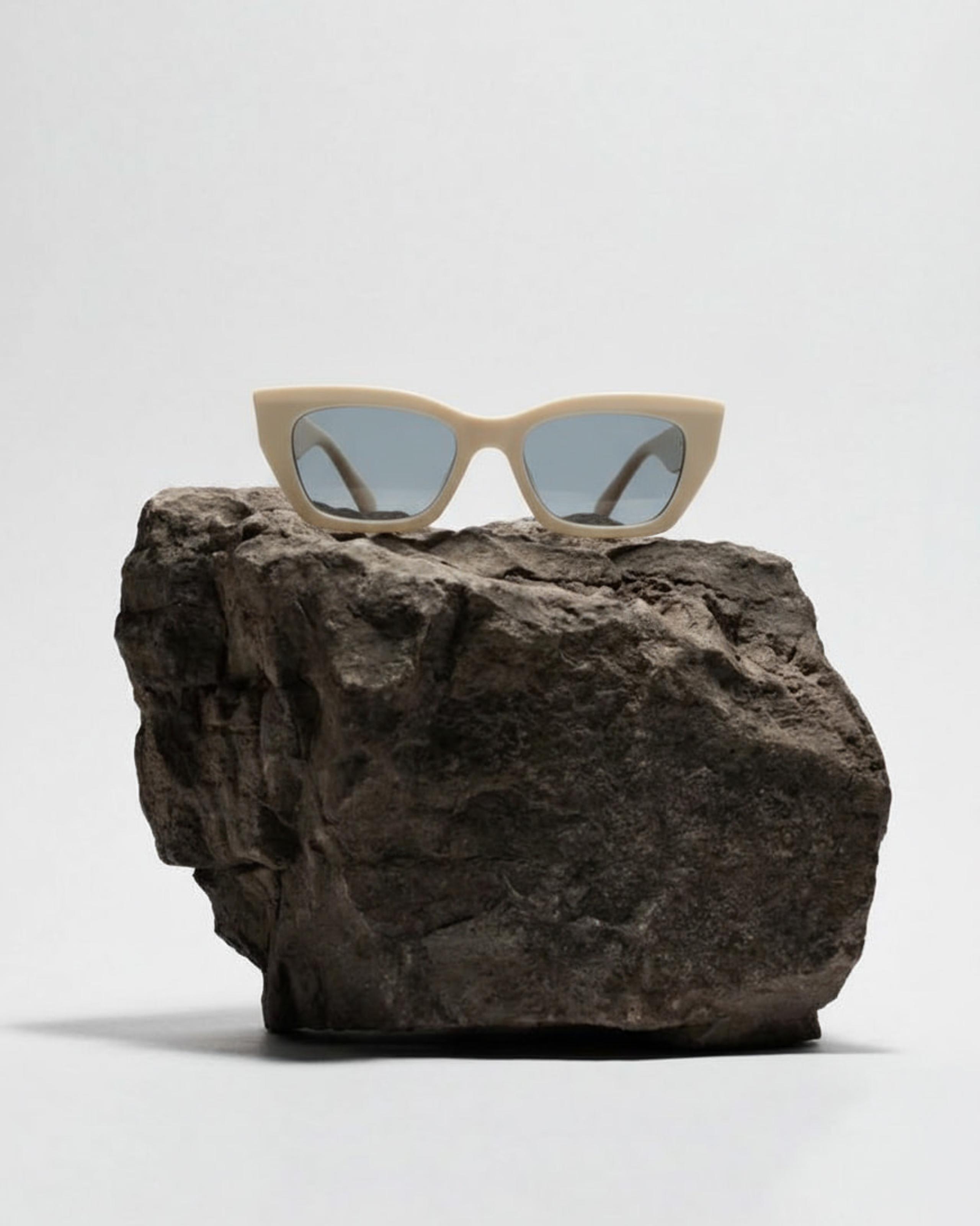 Cream cat-eye sunglasses with tinted lenses perched atop a rough grey stone plinth on grey backdrop