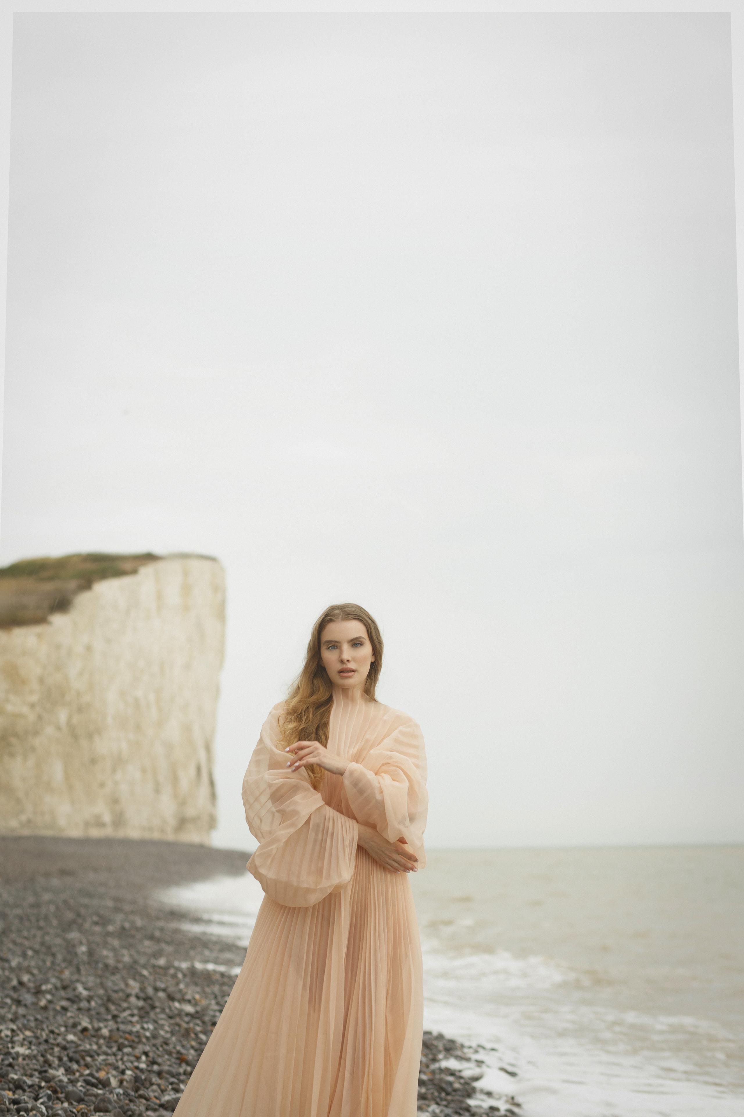 Model in flowing peach pleated gown stands on a pebble beach below a chalk cliff under grey sky