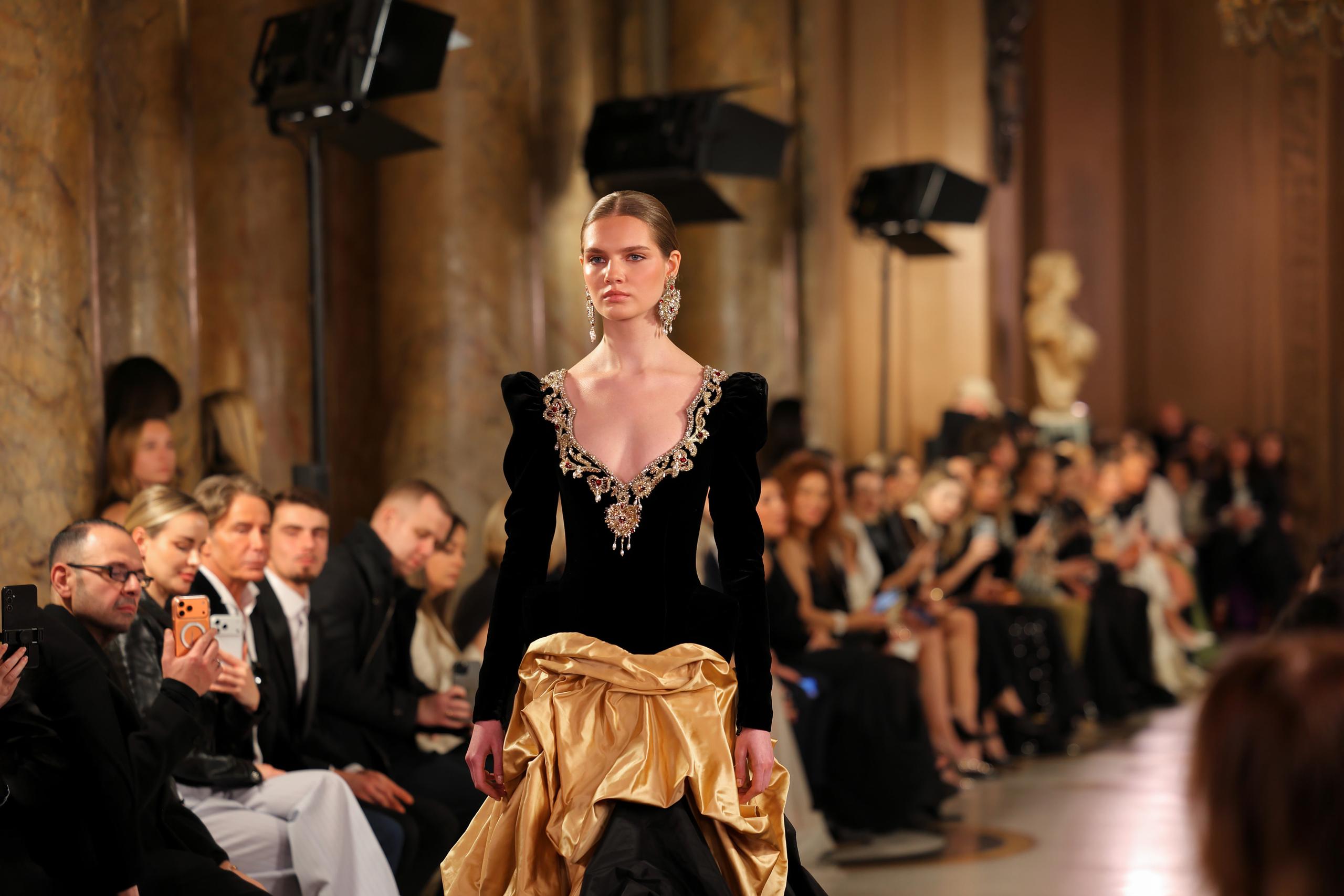 Model in black velvet jeweled bodice gown with gold draped skirt on dim runway