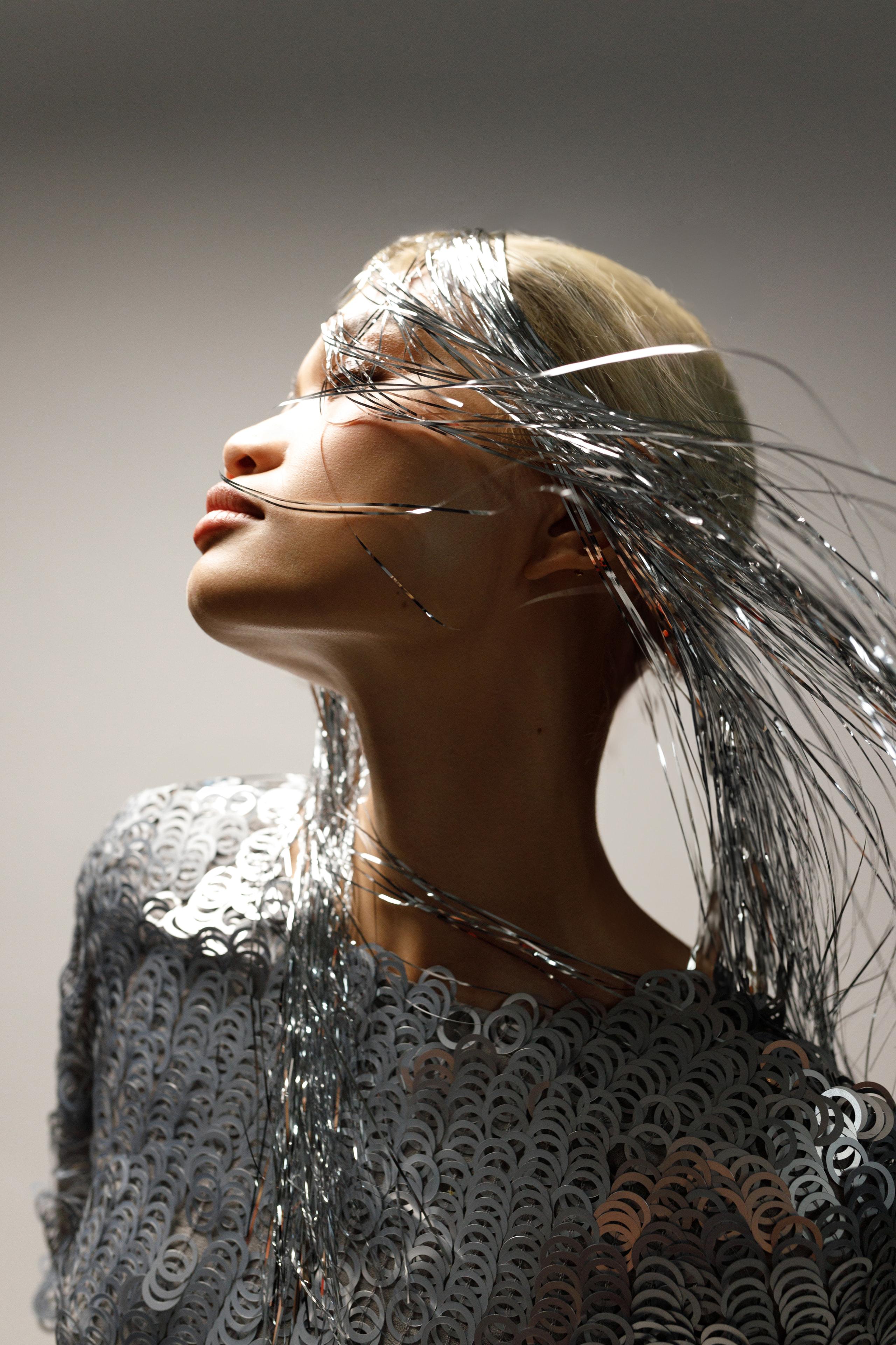 Side profile of model, chin lifted, silver tinsel hair streaming, chrome sequin garment, grey studio.