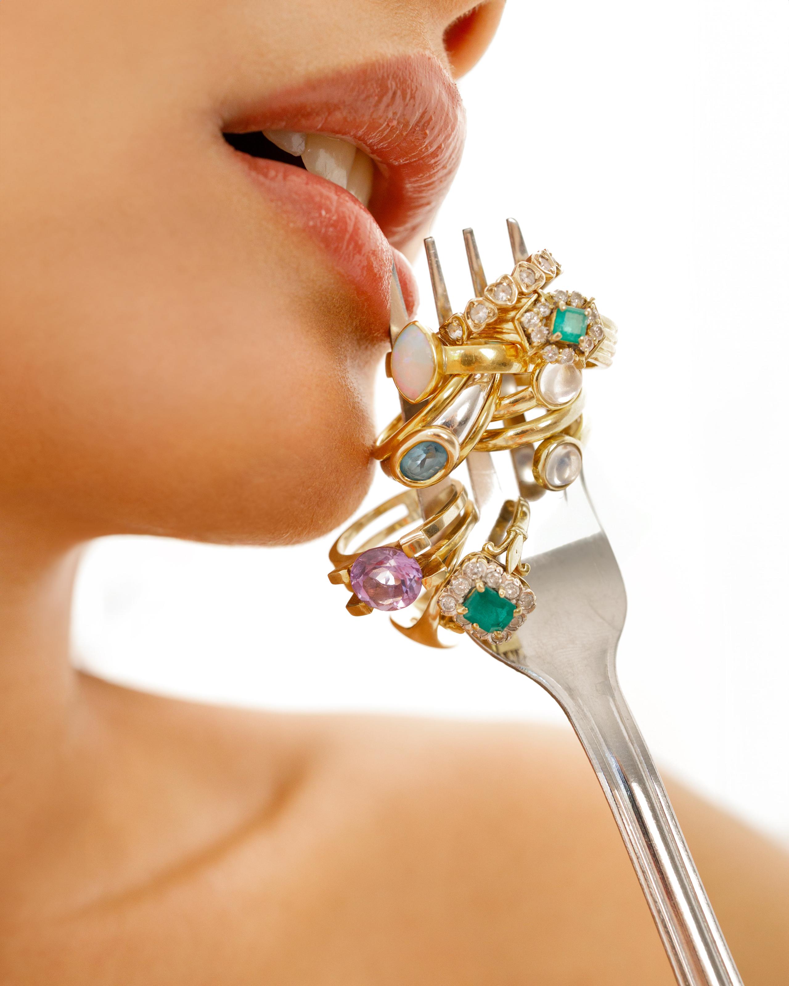 Profile of lips biting a silver fork stacked with colourful gemstone gold rings, bright backdrop
