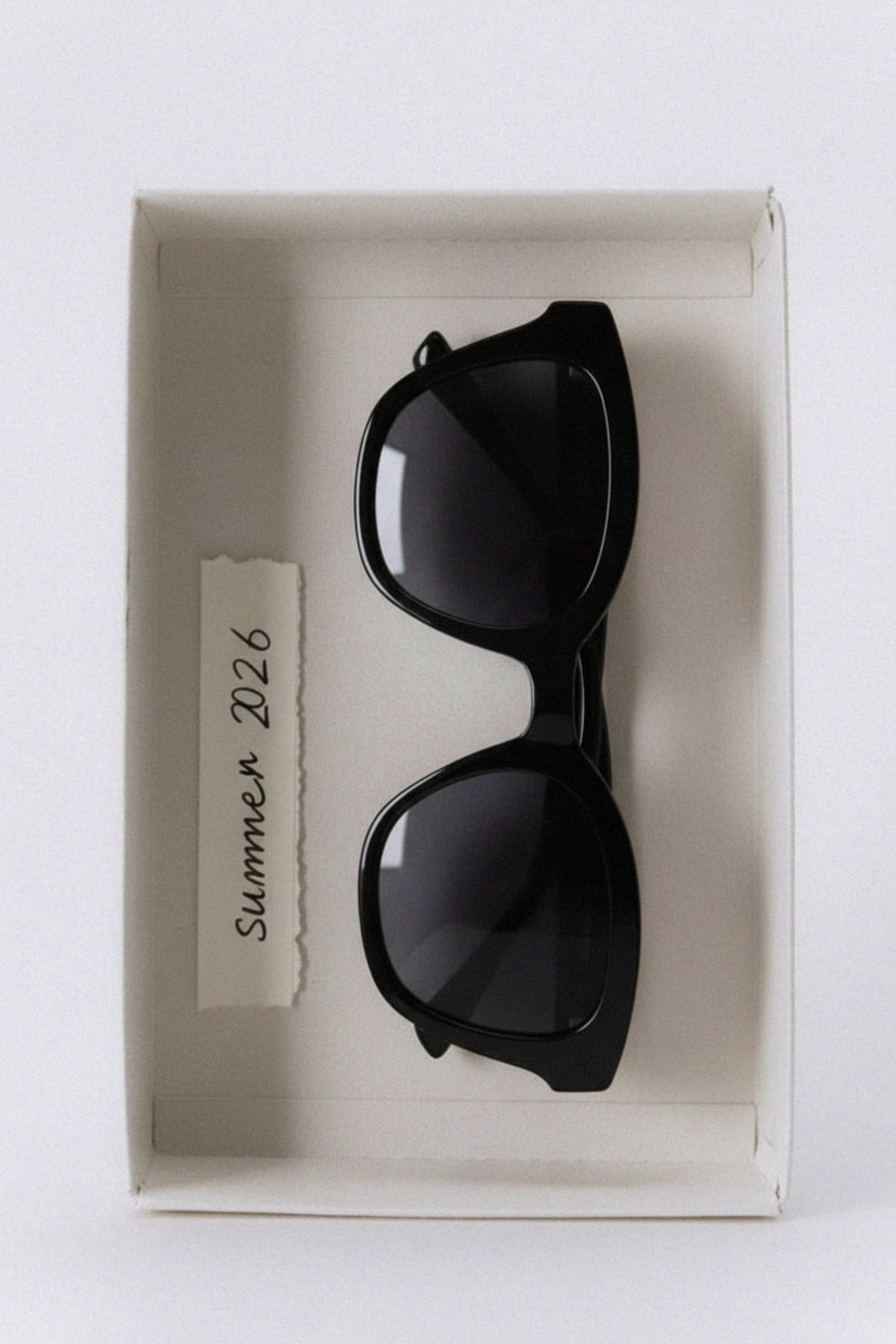 Black wayfarer sunglasses lying in a cream paper box beside a handwritten Summer 2026 label
