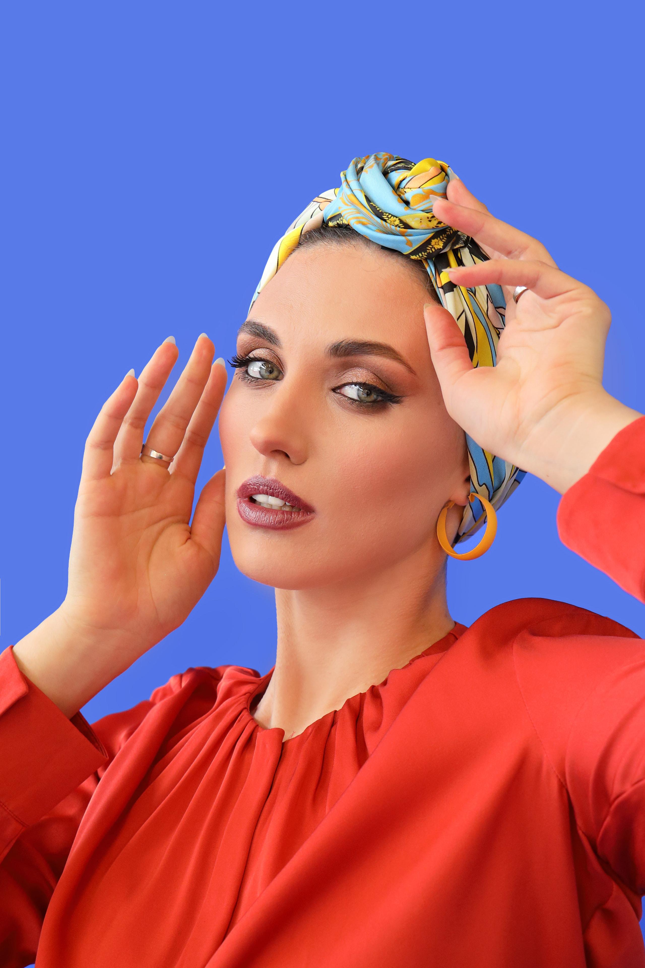 Model in coral blouse with patterned scarf turban gazing past camera on blue