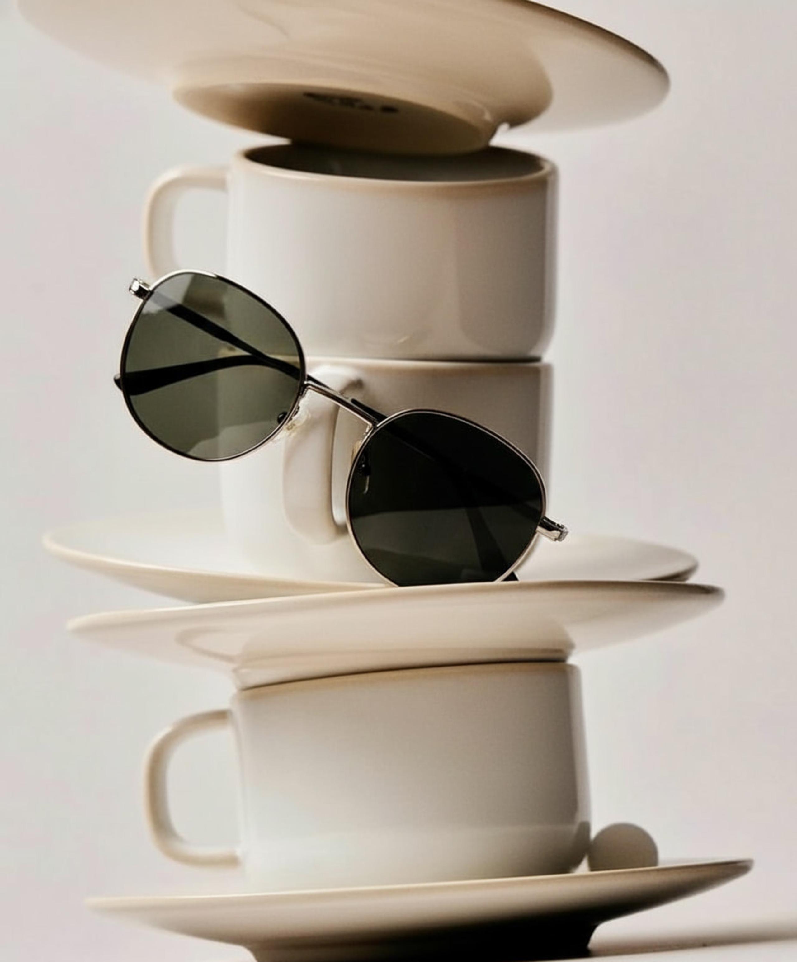 Round silver-framed dark-lens sunglasses balanced on a tilting stack of cream coffee cups and saucers