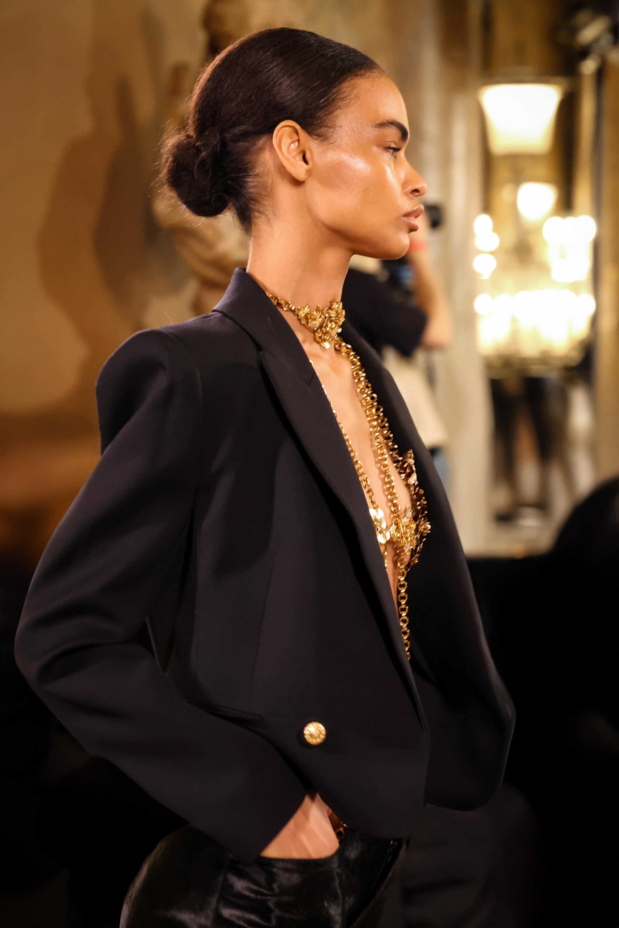 Side profile of model in black blazer draped with layered gold chains, sleek bun, runway lights