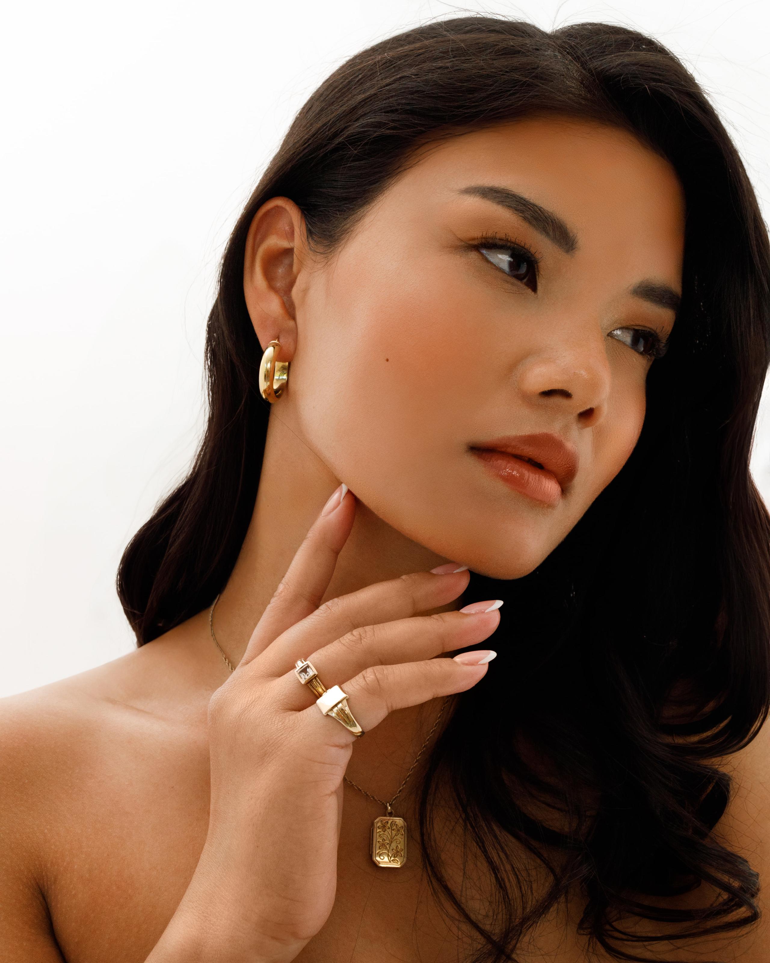 Bare-shouldered model touching her jaw, wearing gold hoop earrings, rings and pendant, soft daylight