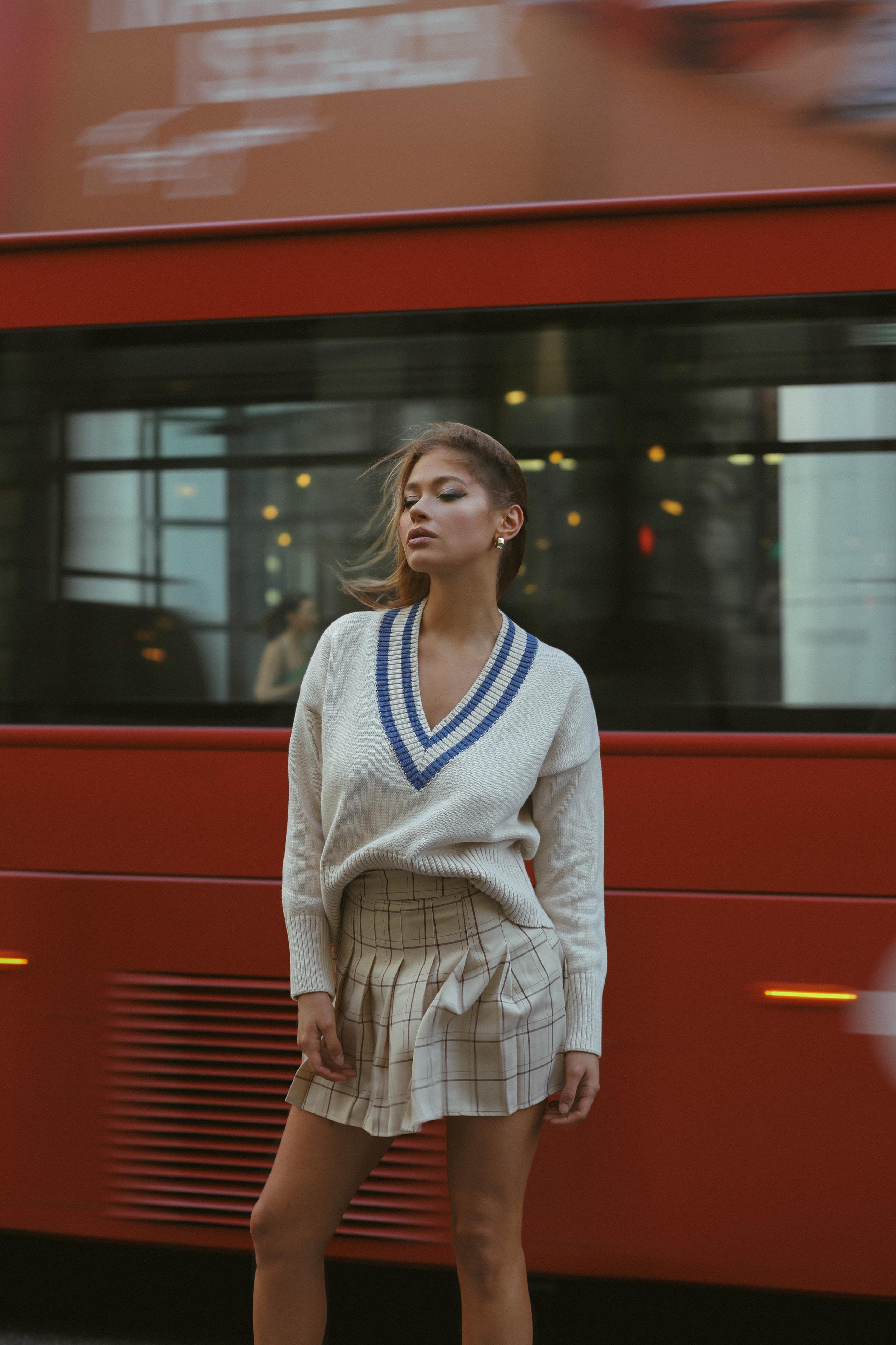 Woman in cream tennis sweater and pleated skirt before a blurred red London bus