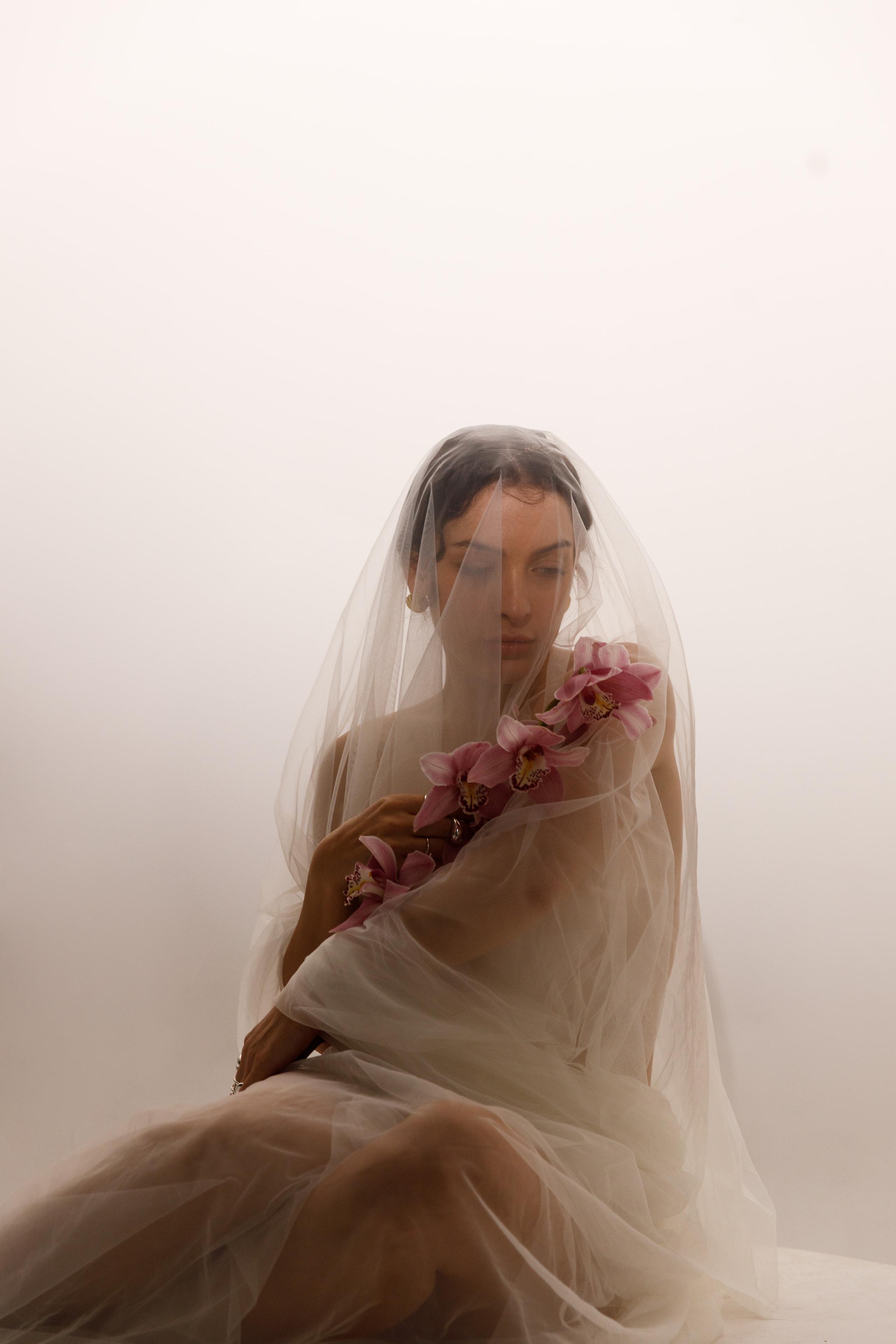 Woman seated draped in sheer white veil holding pink orchids, dissolving into high-key white haze