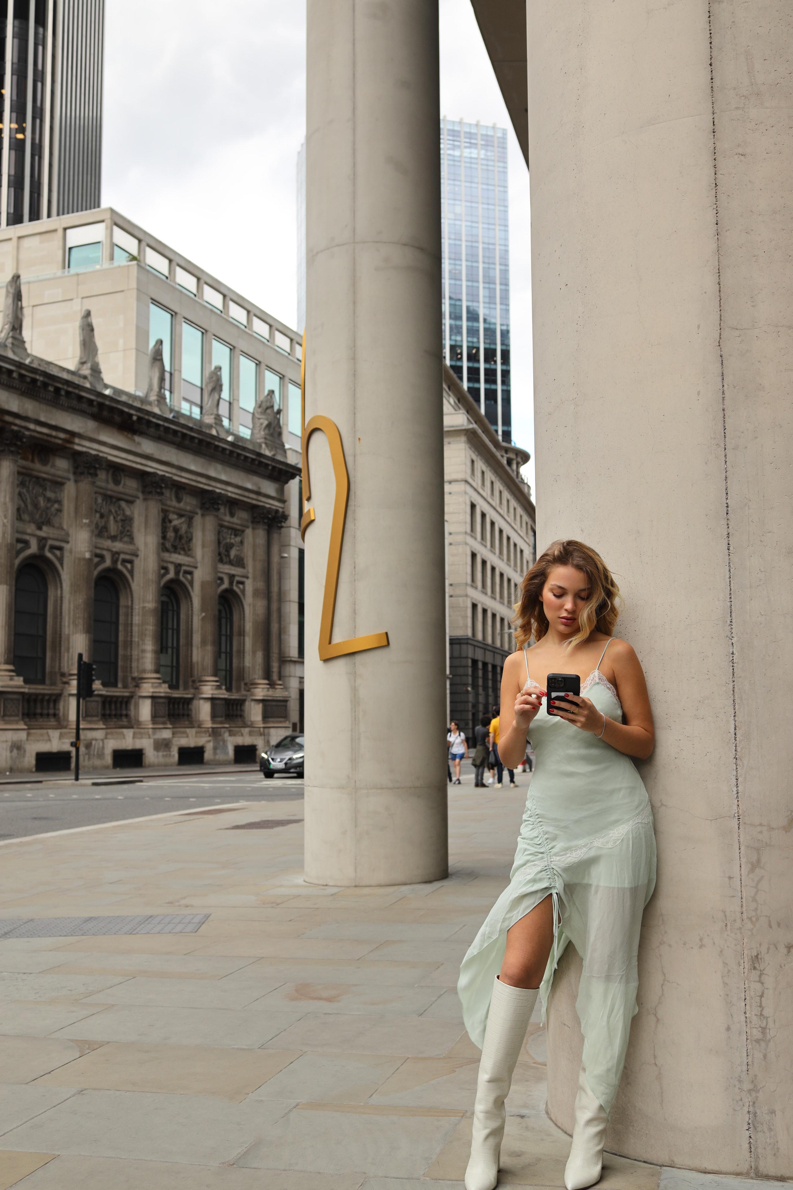 Woman in sage slip dress and white boots leaning on column, checking phone, London financial district