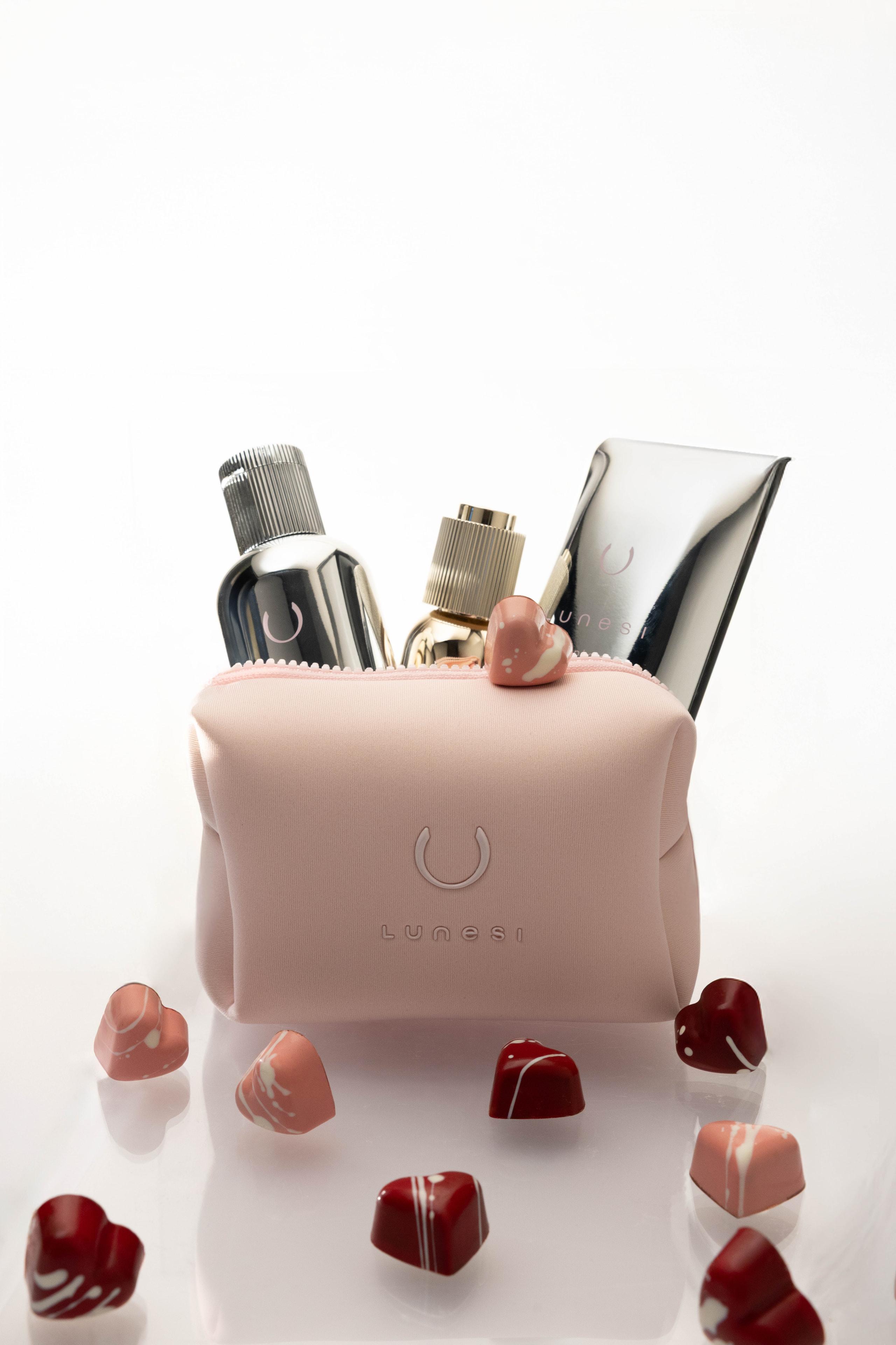 Blush pink Lunesi pouch holding chrome and gold products surrounded by chocolate hearts on white