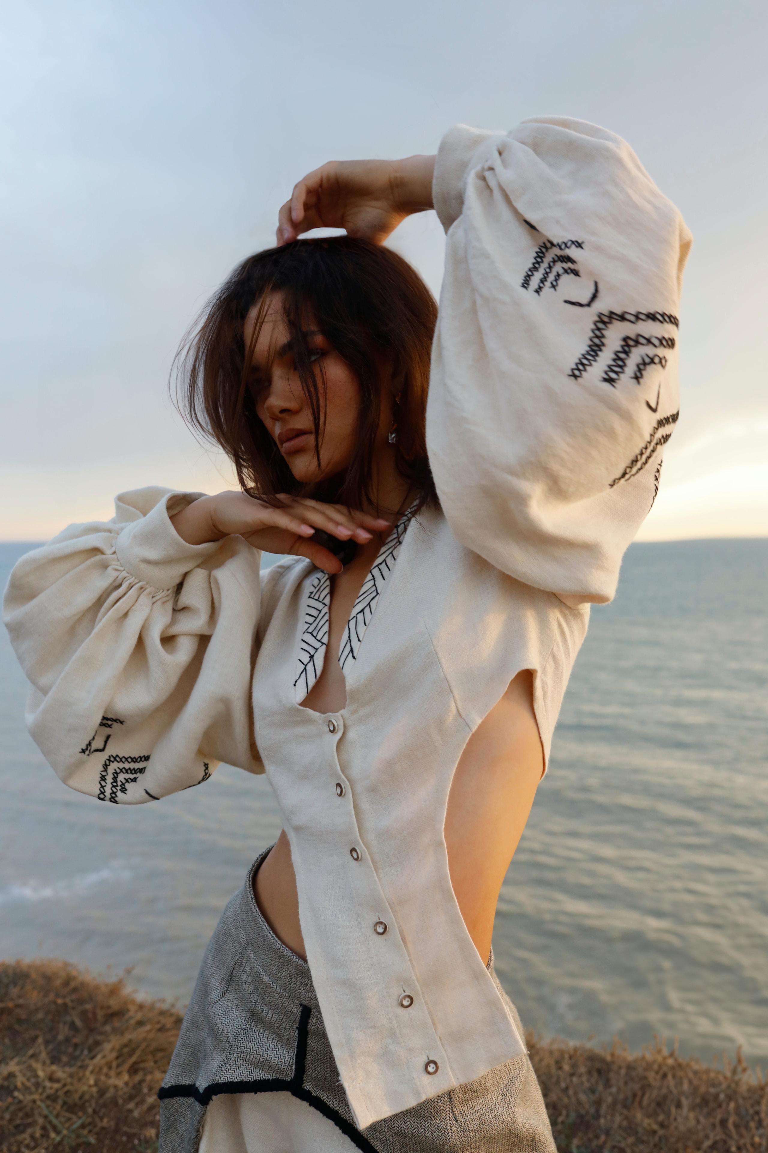Woman in cream embroidered linen blouse with puffed sleeves posing on grassy clifftop above the sea
