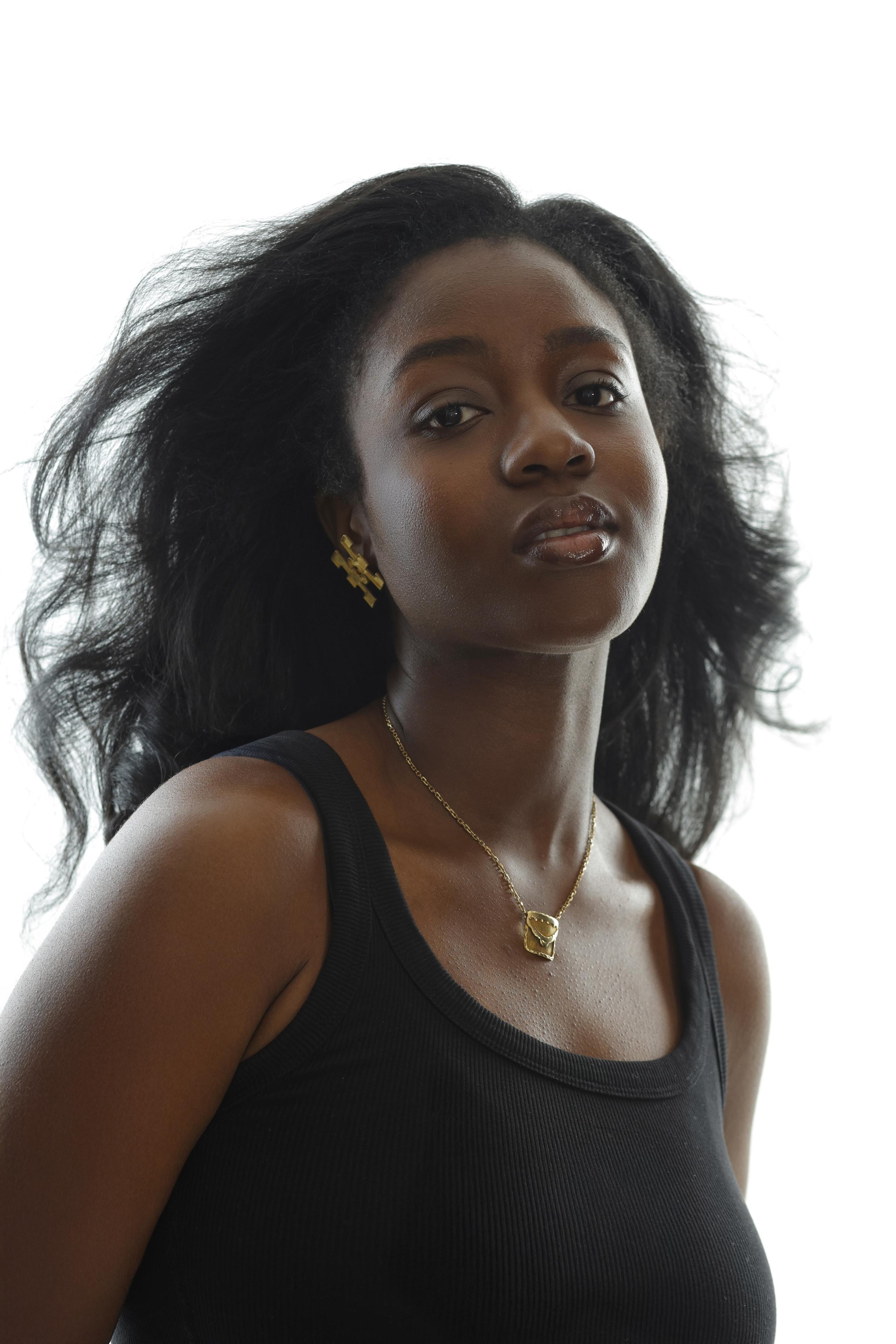 Woman in a black tank top with voluminous wind-swept hair gazes confidently against a white studio