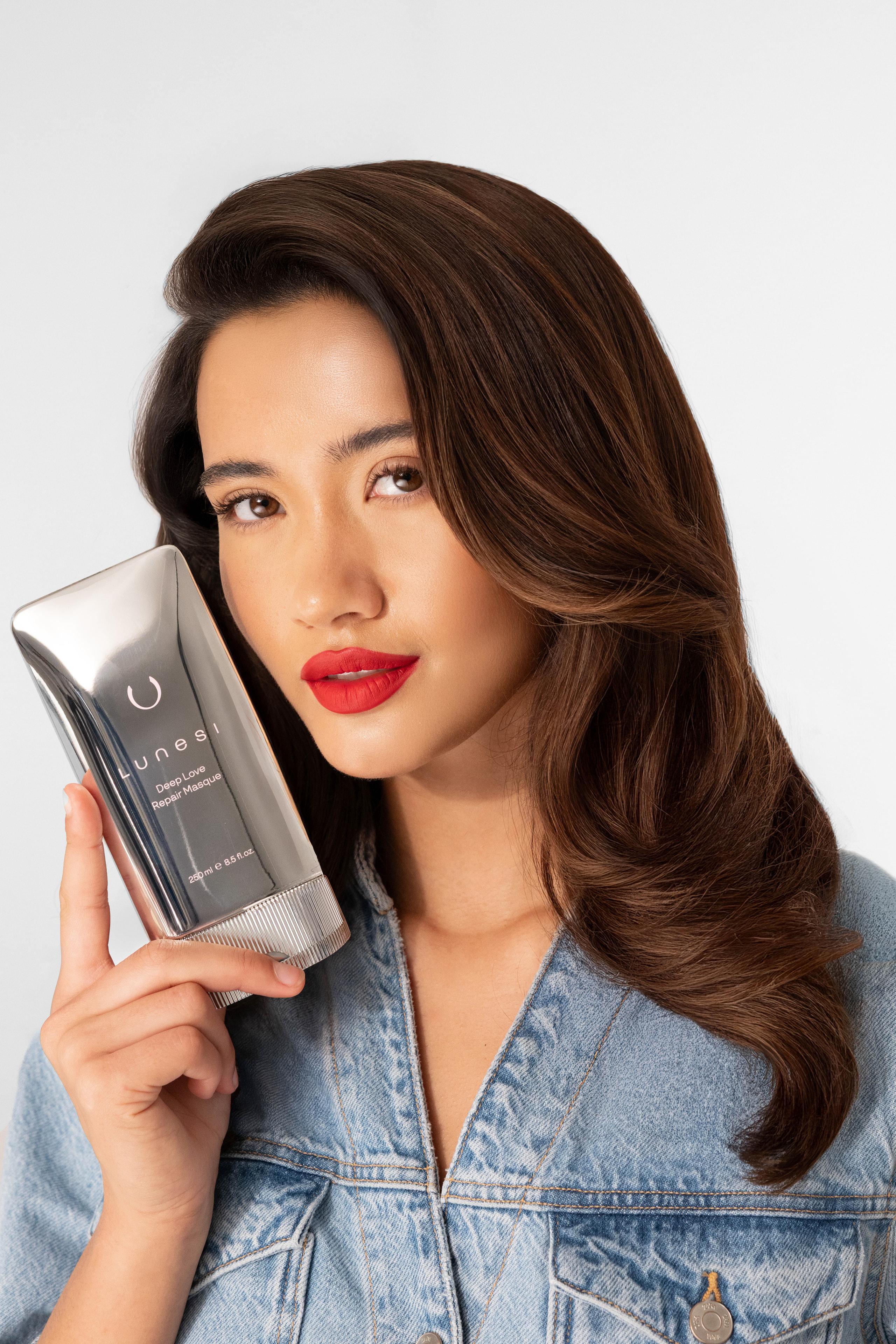 Model with voluminous wavy hair holds a chrome Lunesi Deep Love Repair Masque tube to her cheek