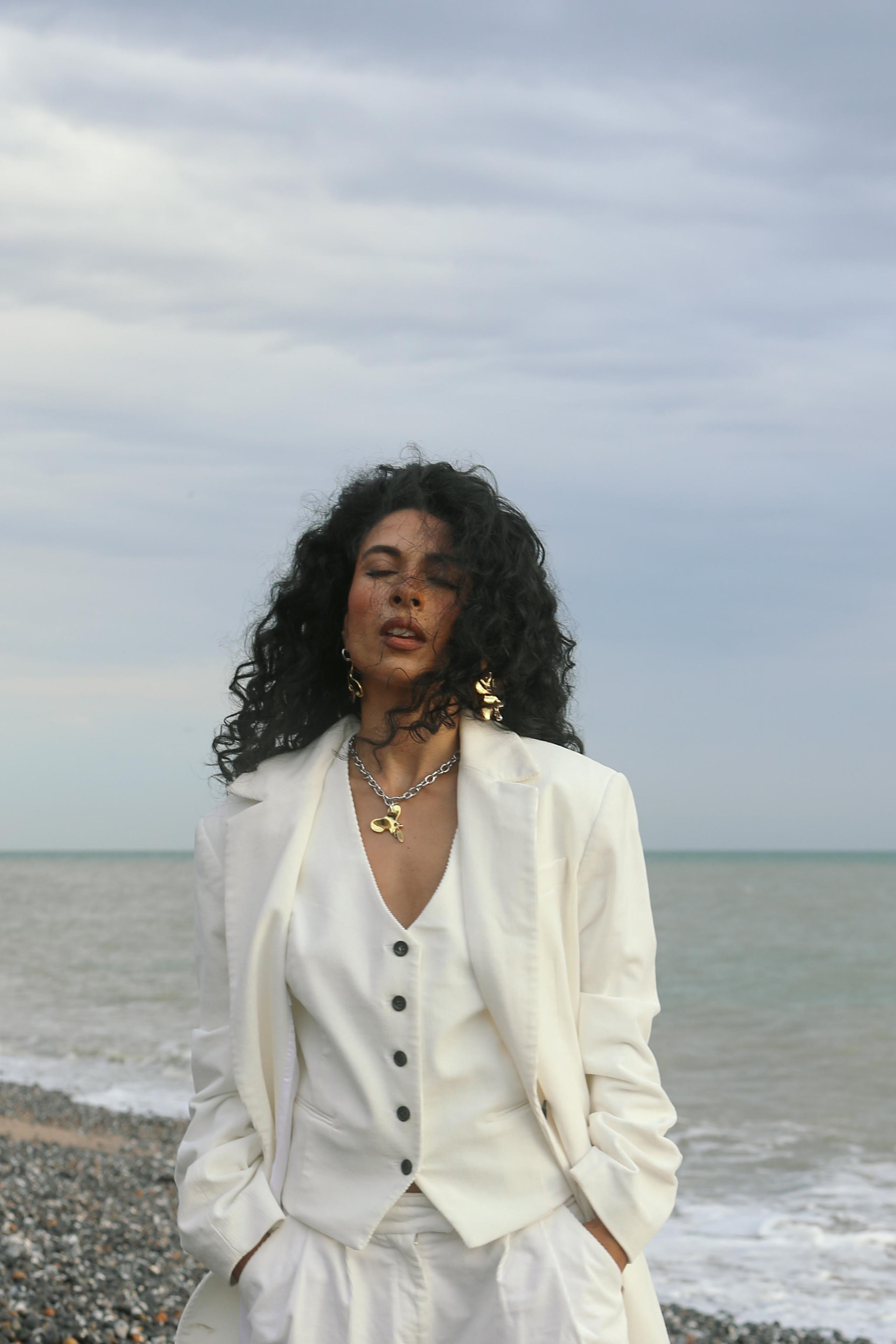 Woman with curly dark hair and closed eyes in cream suit, hands in pockets, calm grey seascape behind