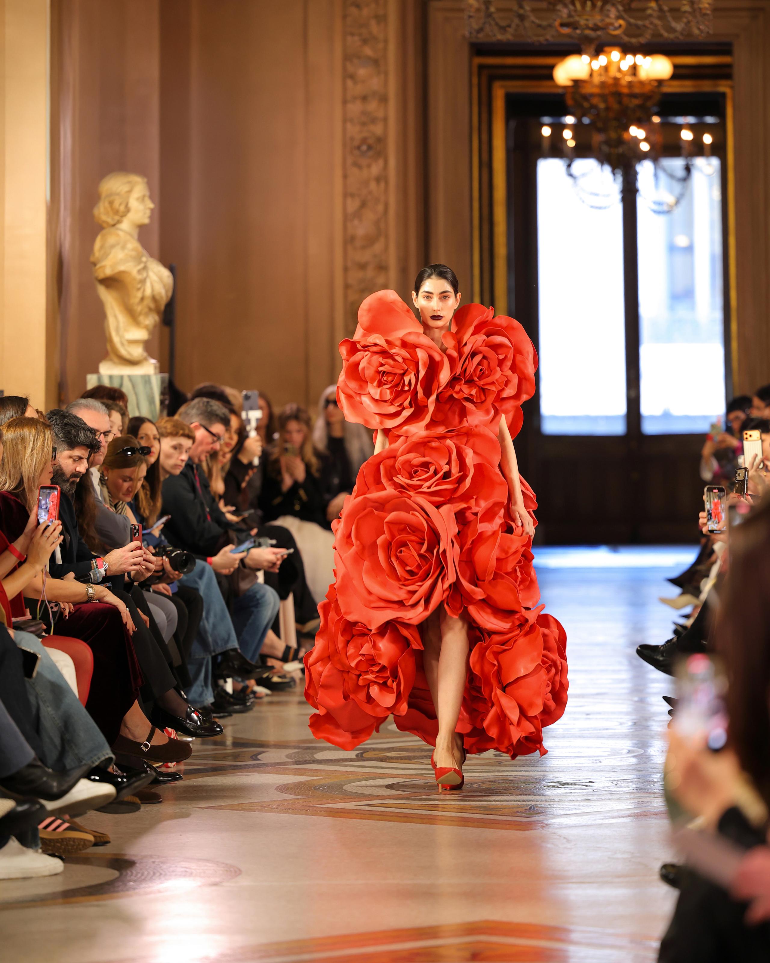 Model in dramatic red dress built from oversized fabric roses walking grand hall runway
