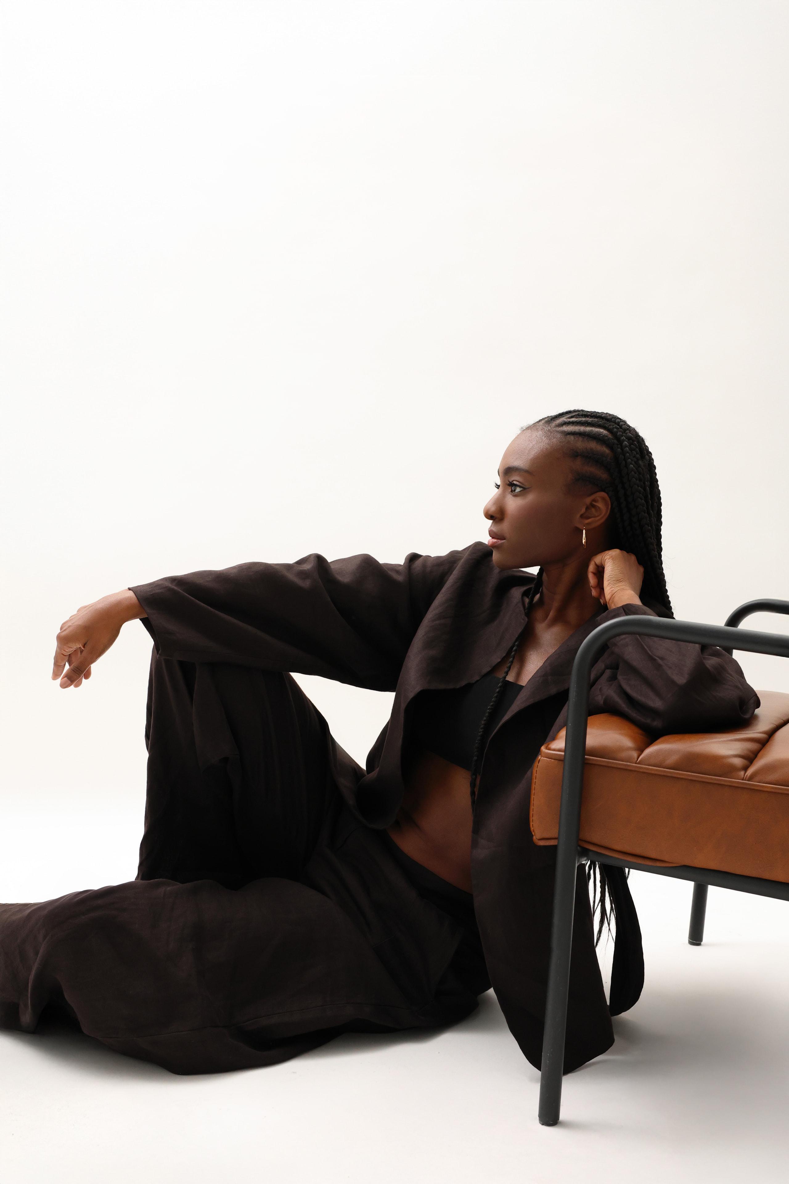 Woman in brown linen suit sits on floor by leather bench, gazing aside in bright studio