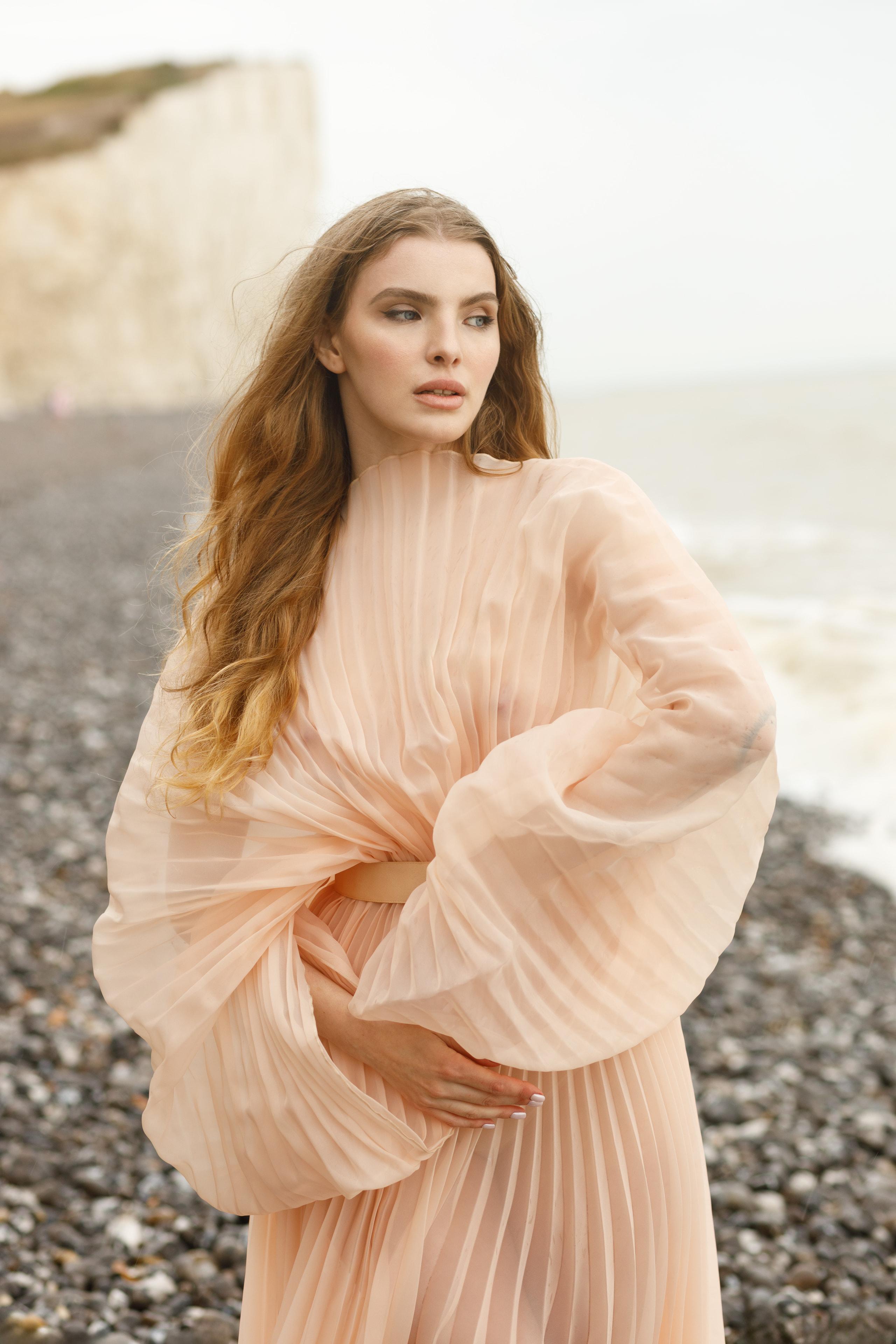 Three-quarter portrait of woman with long wavy hair in peach pleated gown on windswept pebble beach