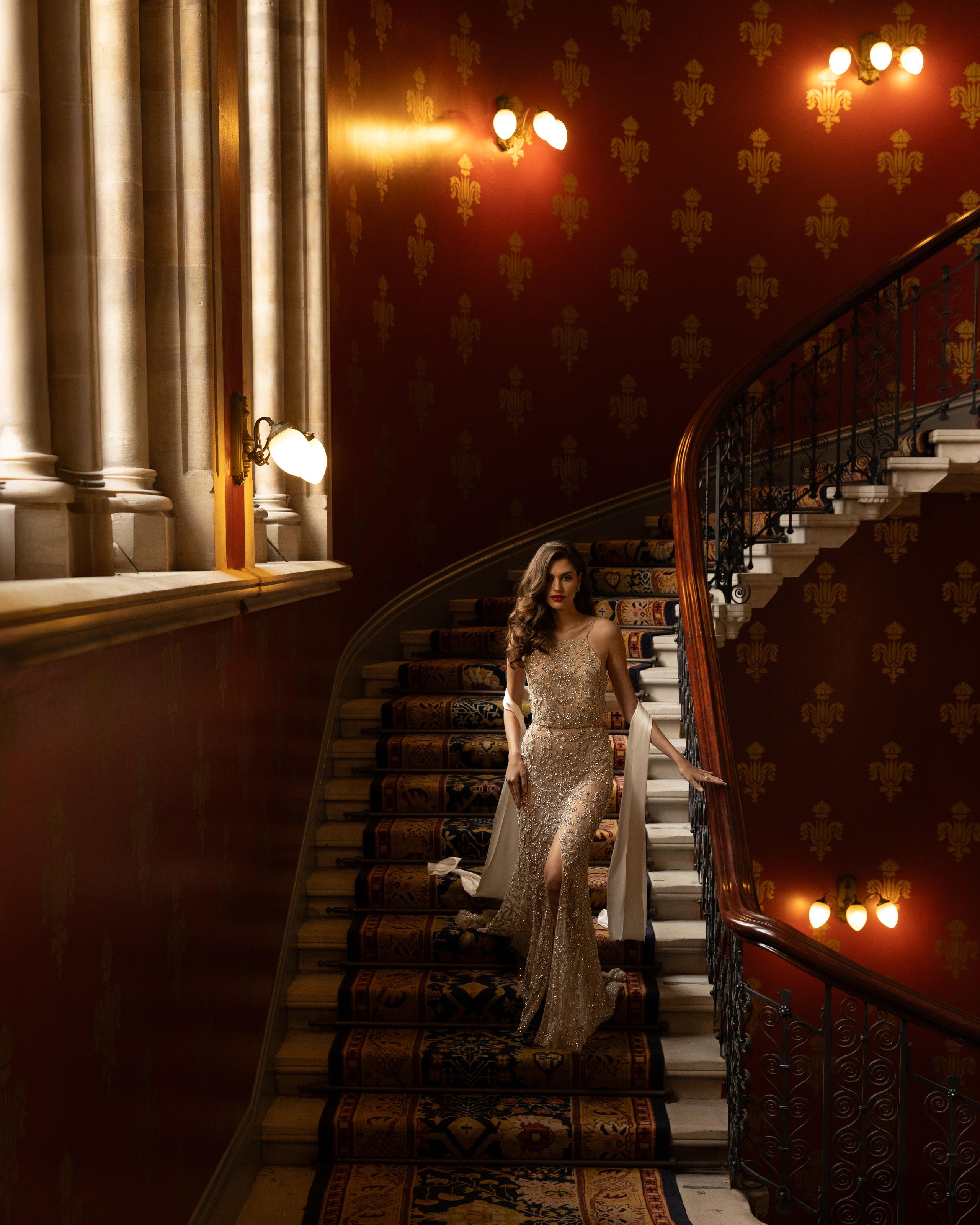 Model in beaded gown on grand carpeted staircase, stone columns, lit sconces, red damask walls