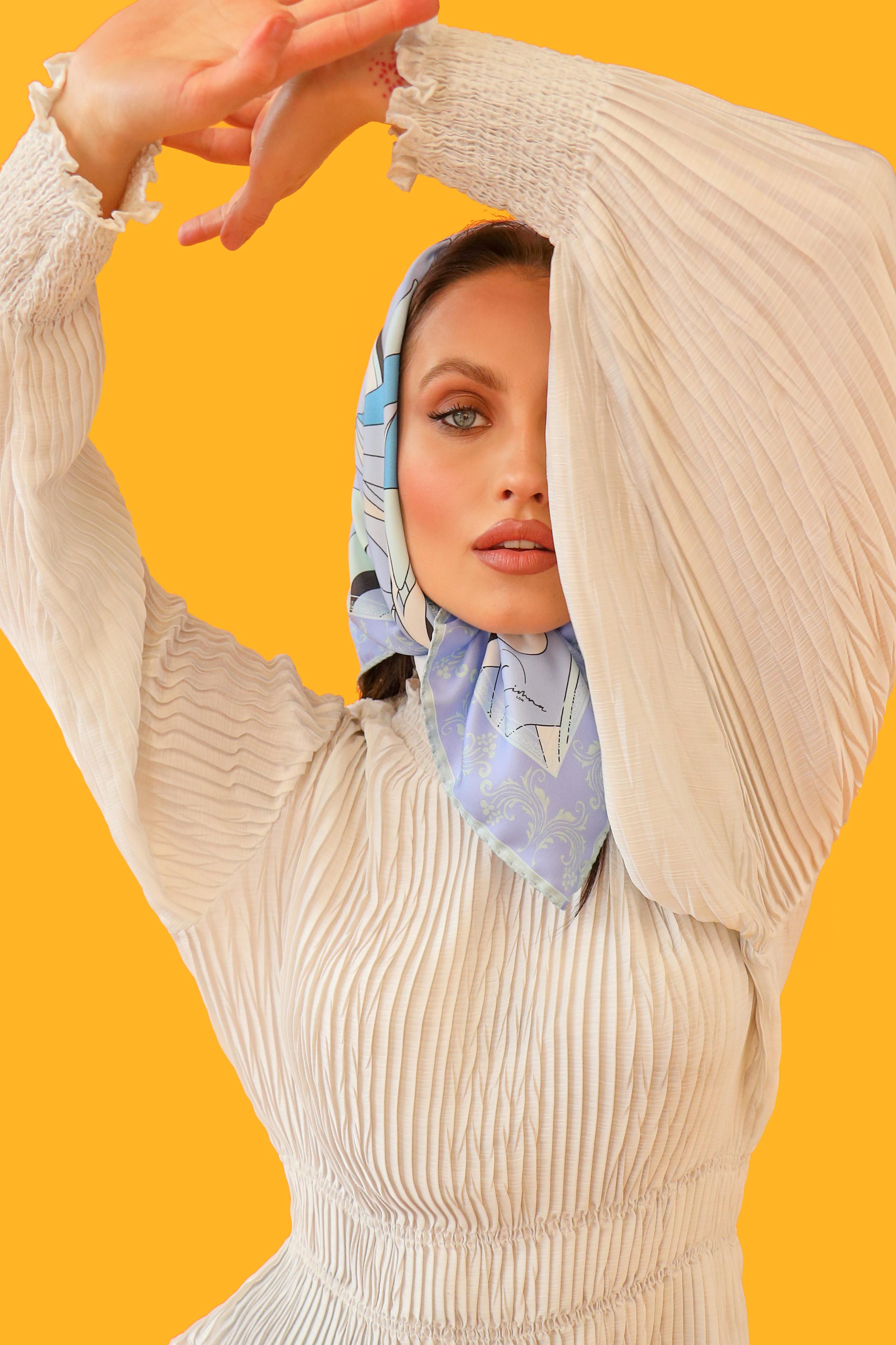 Model in pleated cream dress and pale blue scarf raising arms overhead frame