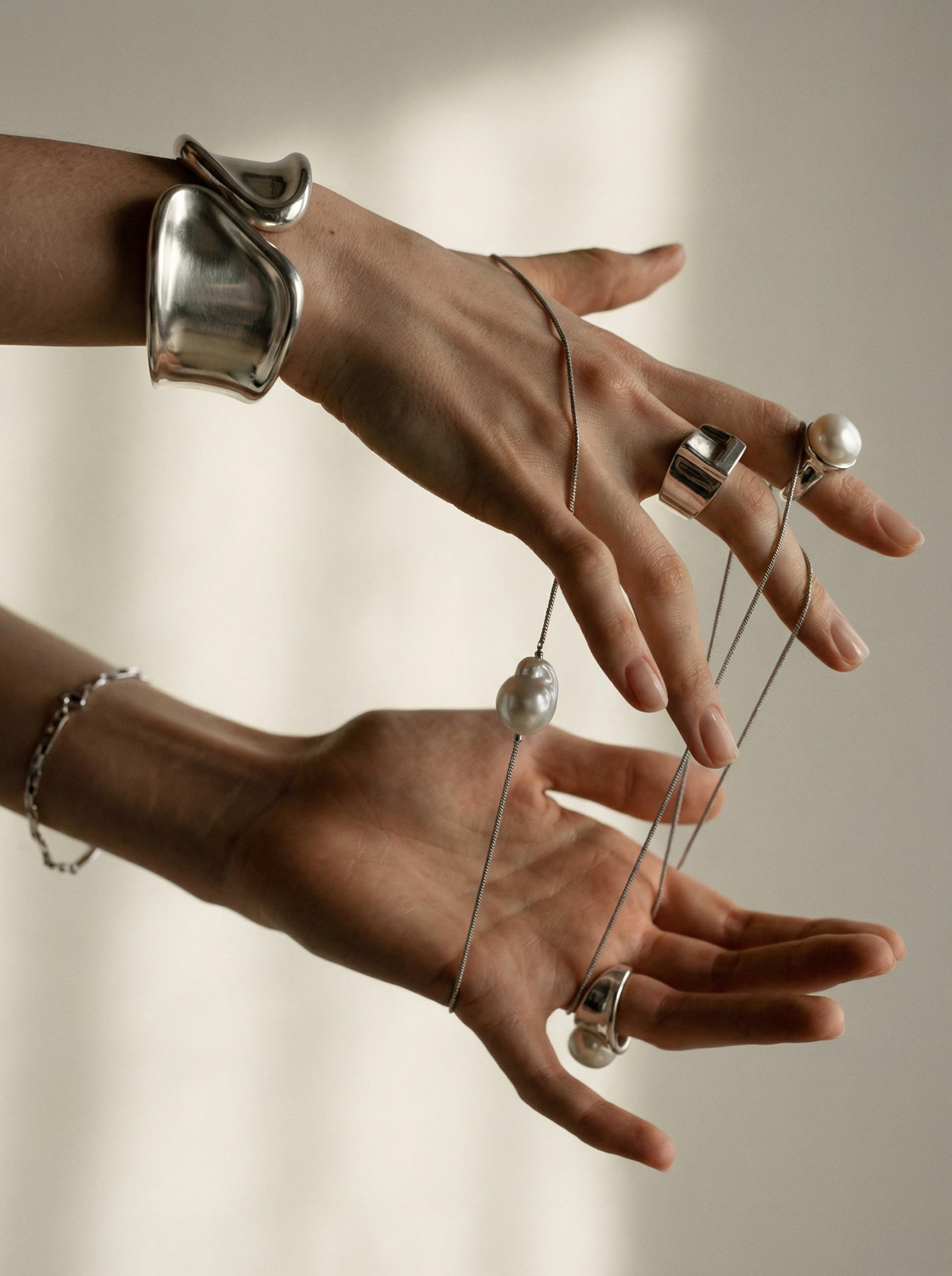 Two hands modelling silver cuff, chunky rings, pearl pendant and chain bracelet