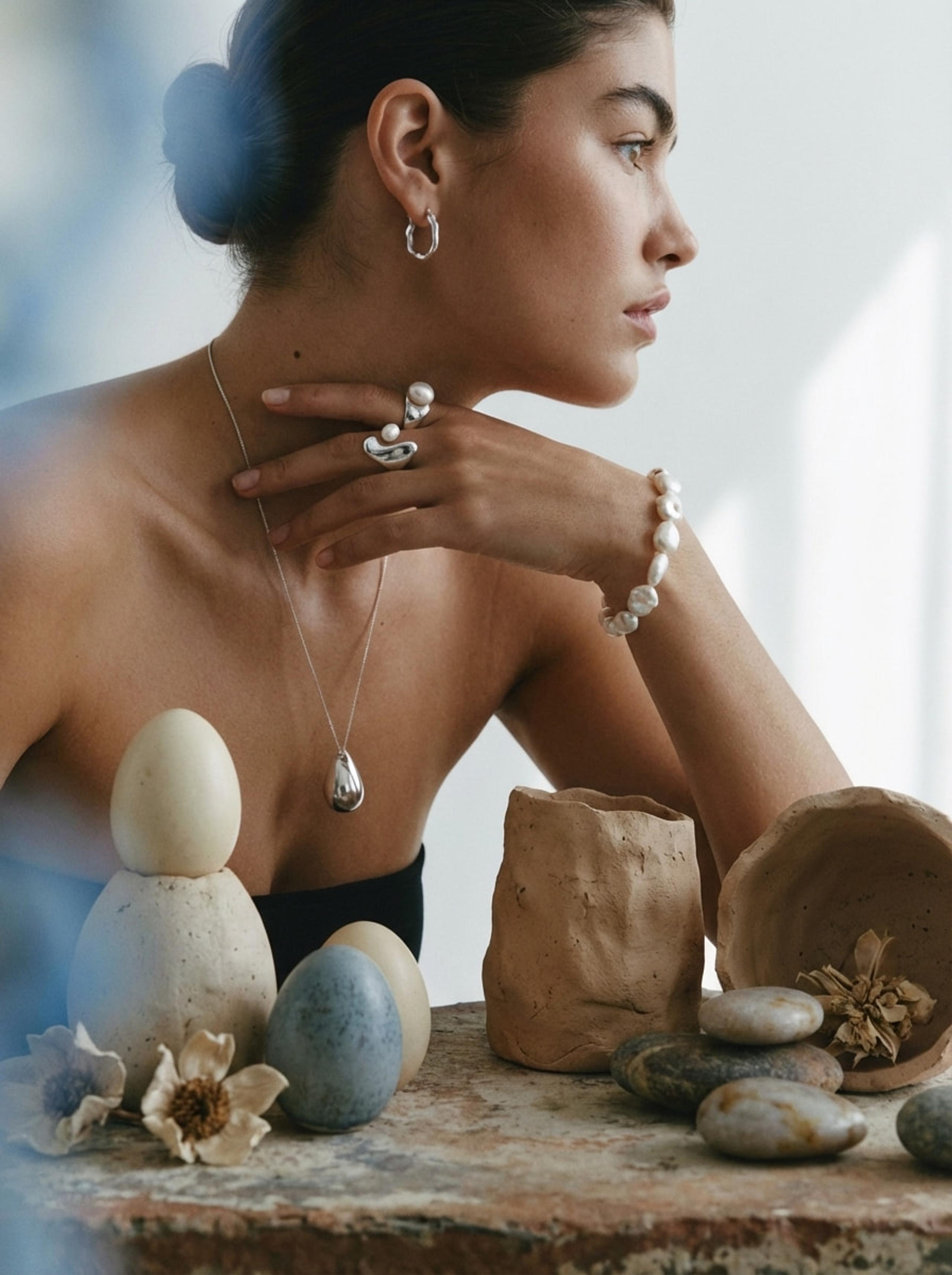 Woman in profile wearing silver and pearl jewellery beside clay vessels, stones and dried flowers, soft light.