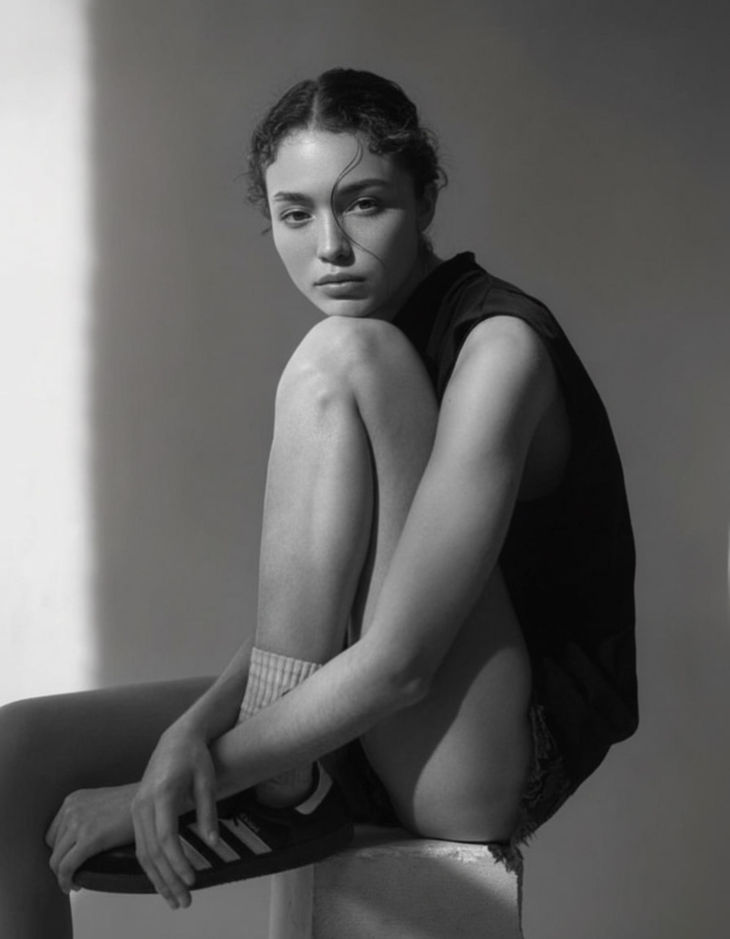 Monochrome portrait of woman seated with knees up, holding sneaker, direct gaze, soft shadow wall