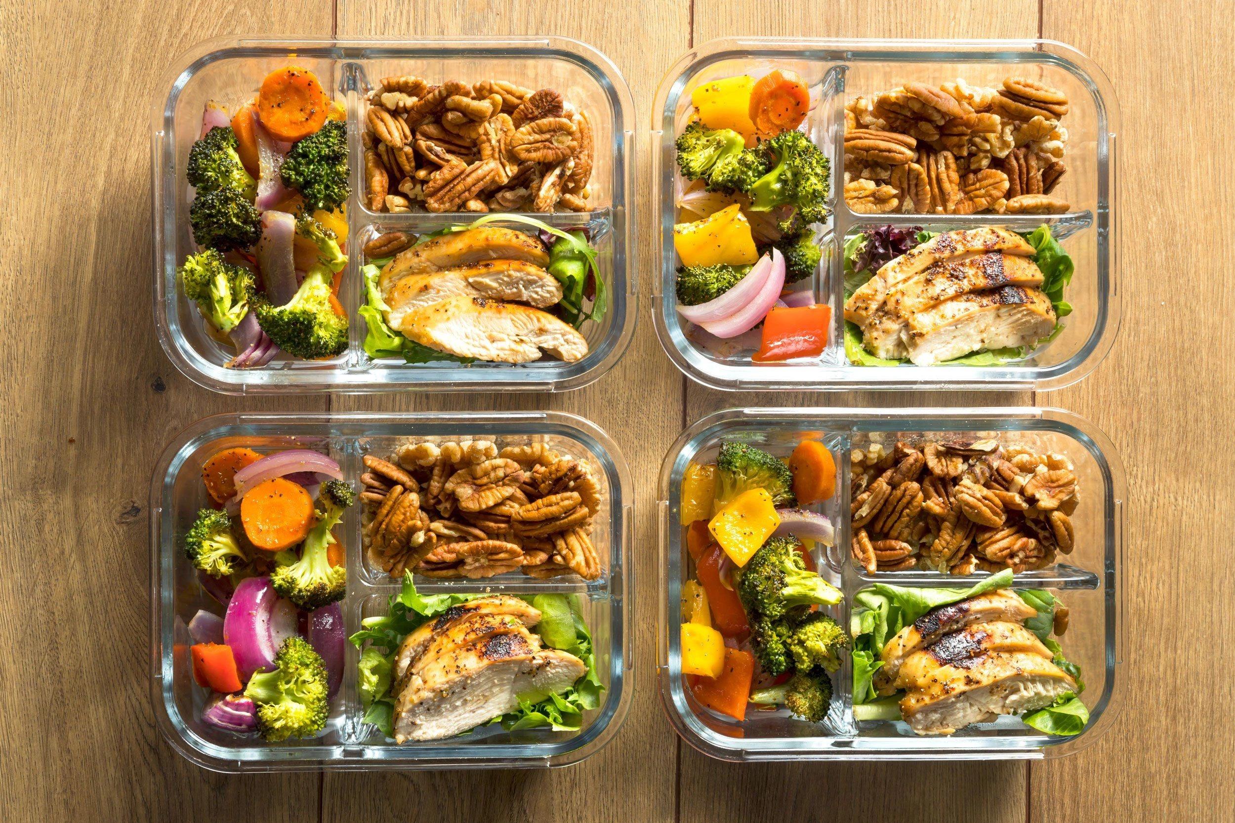 Meal prep containers filled with grilled chicken breast, roasted broccoli, carrots, bell peppers, red onions, leafy greens, and mixed nuts arranged neatly on a wooden table. Top-down view with natural lighting and a healthy eating concept.